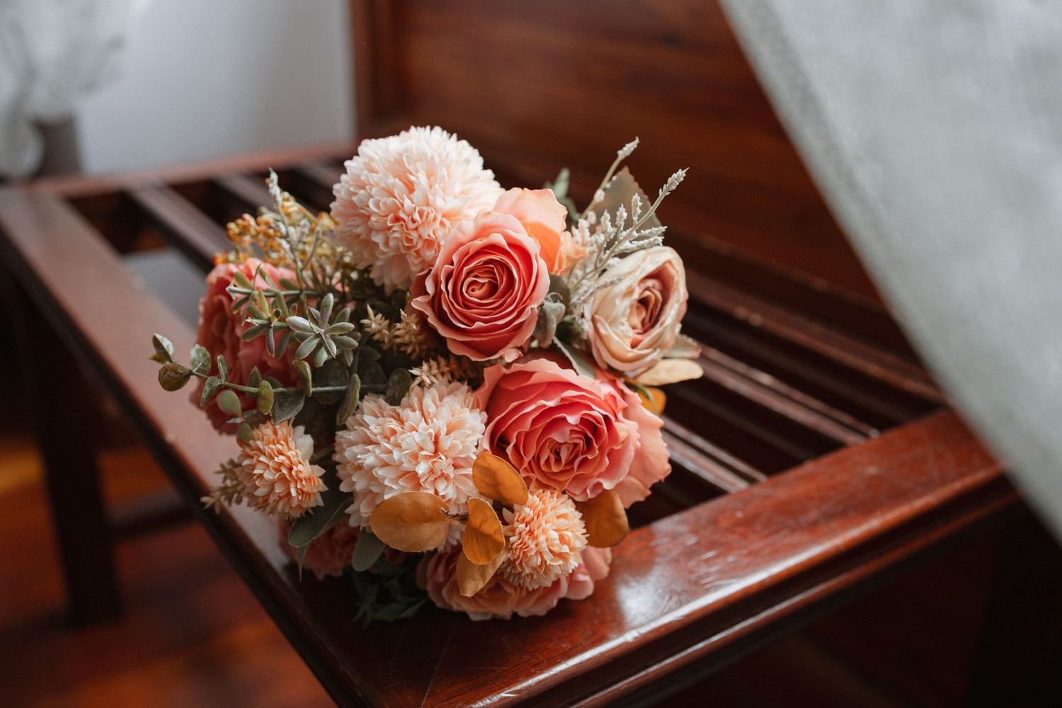 Peach and coral floral arrangement with roses and dried flowers rests on wooden furniture surface in warm lighting.