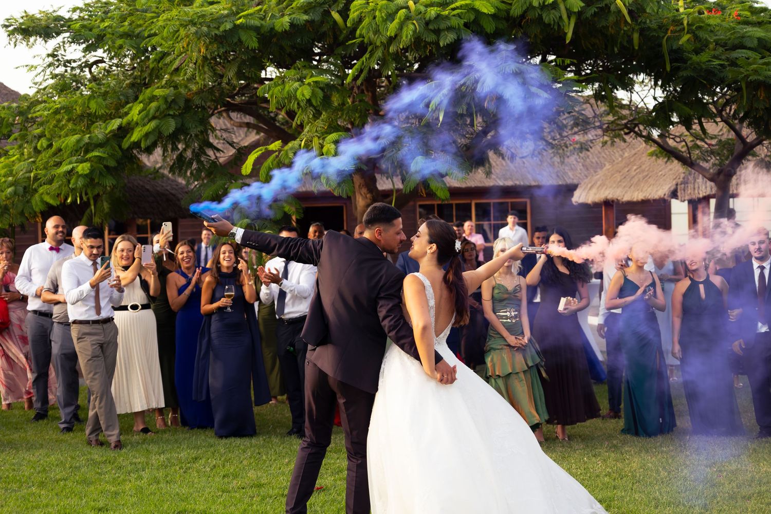Outdoor wedding reception with guests celebrating under trees and blue smoke effects.