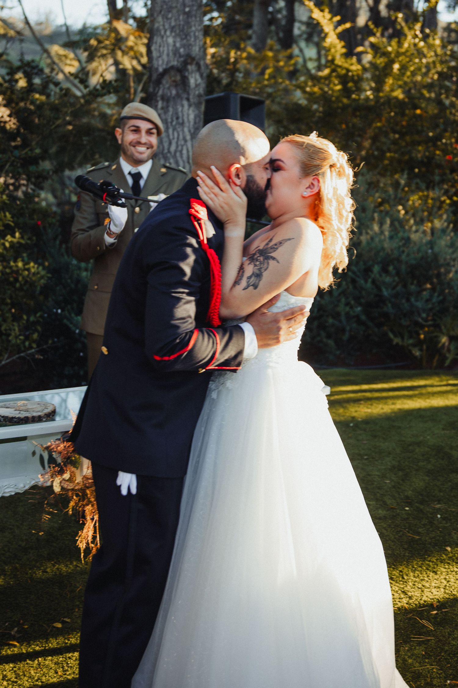 A romantic wedding kiss between newlyweds in formal attire while someone watches in the background outdoors.