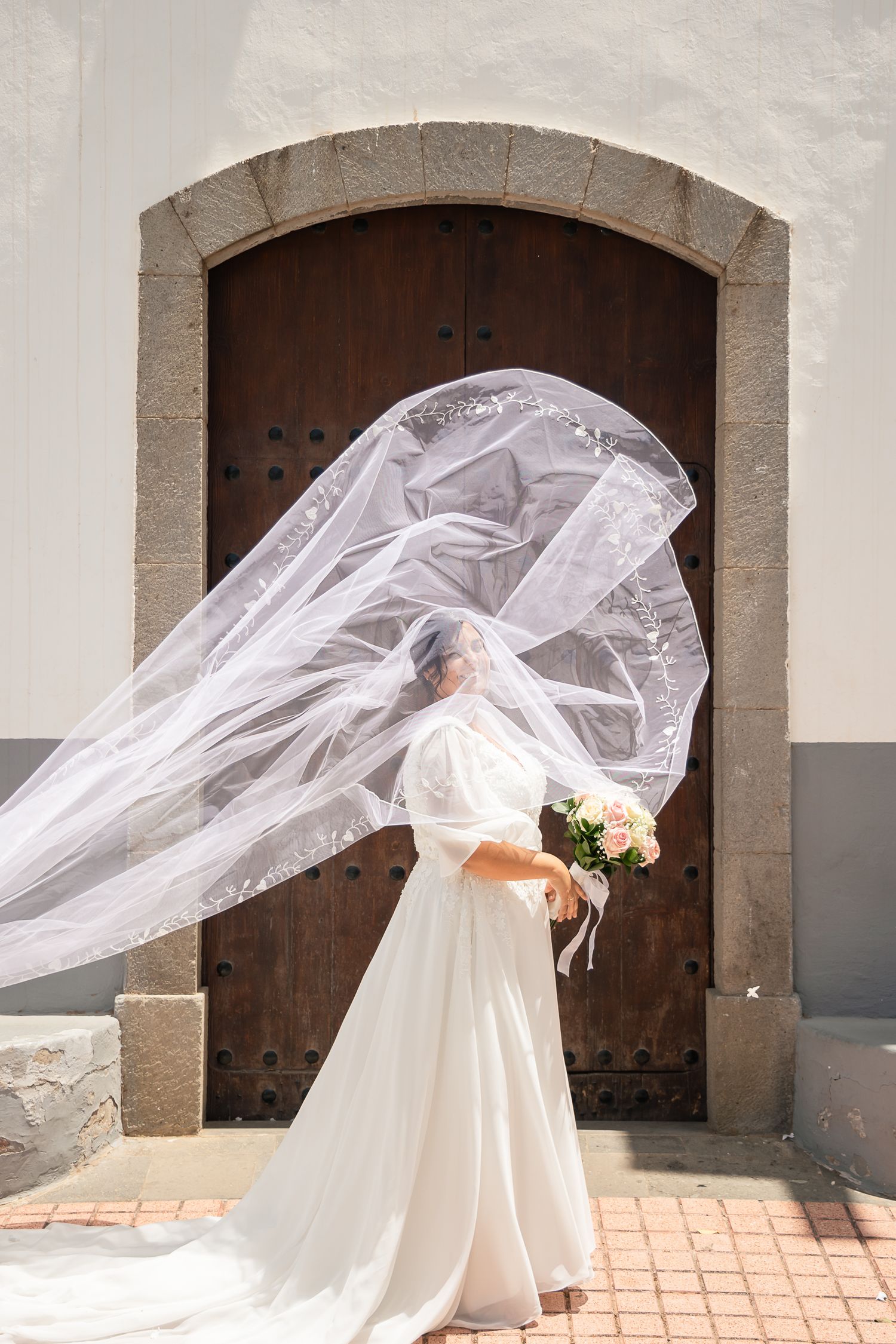 A dramatic veil billows in the wind against stone architecture and wooden doors.