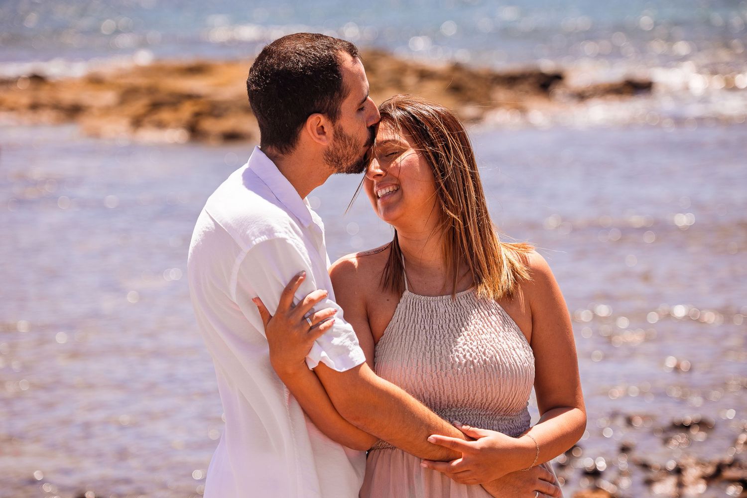 Couple shares intimate moments on a sunny beach with waves in the background.