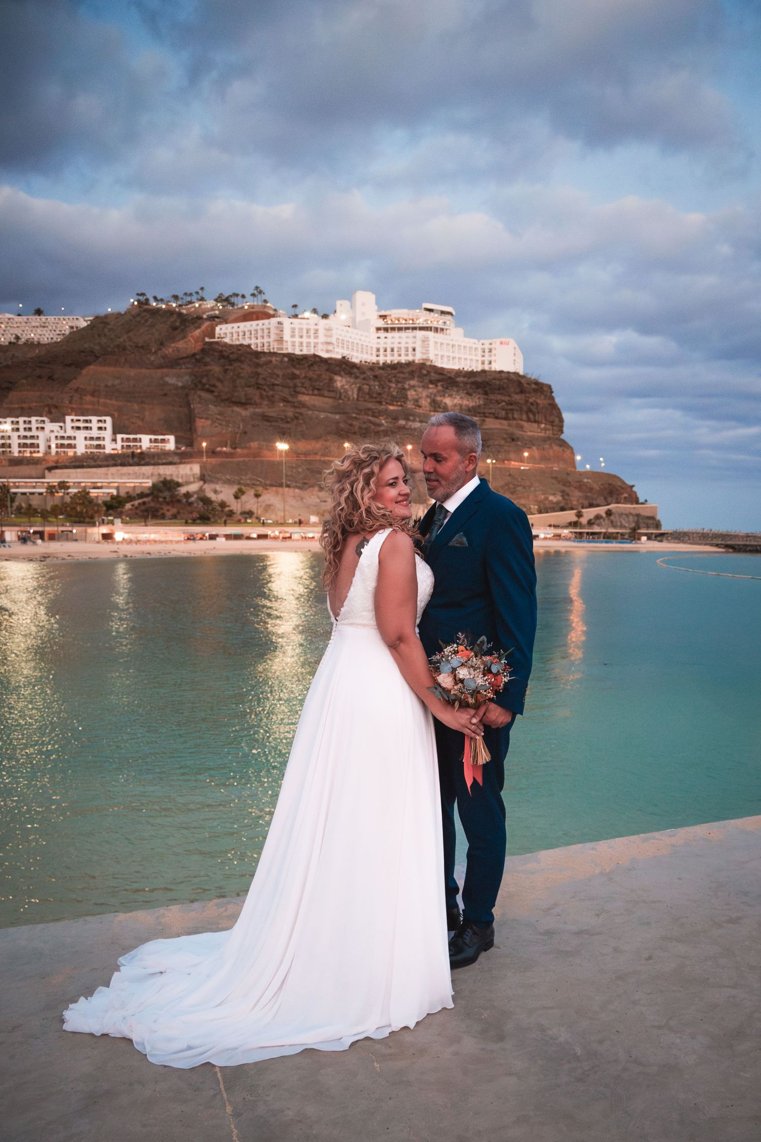 A coastal wedding portrait with turquoise waters and a white cliffside resort in the background.