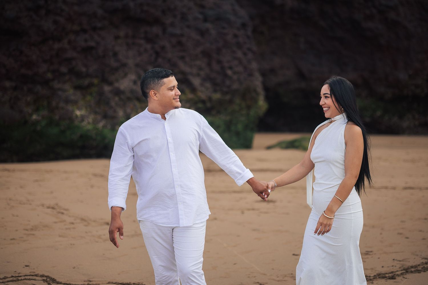 Couple in white outfits walking along sandy beach at sunset.