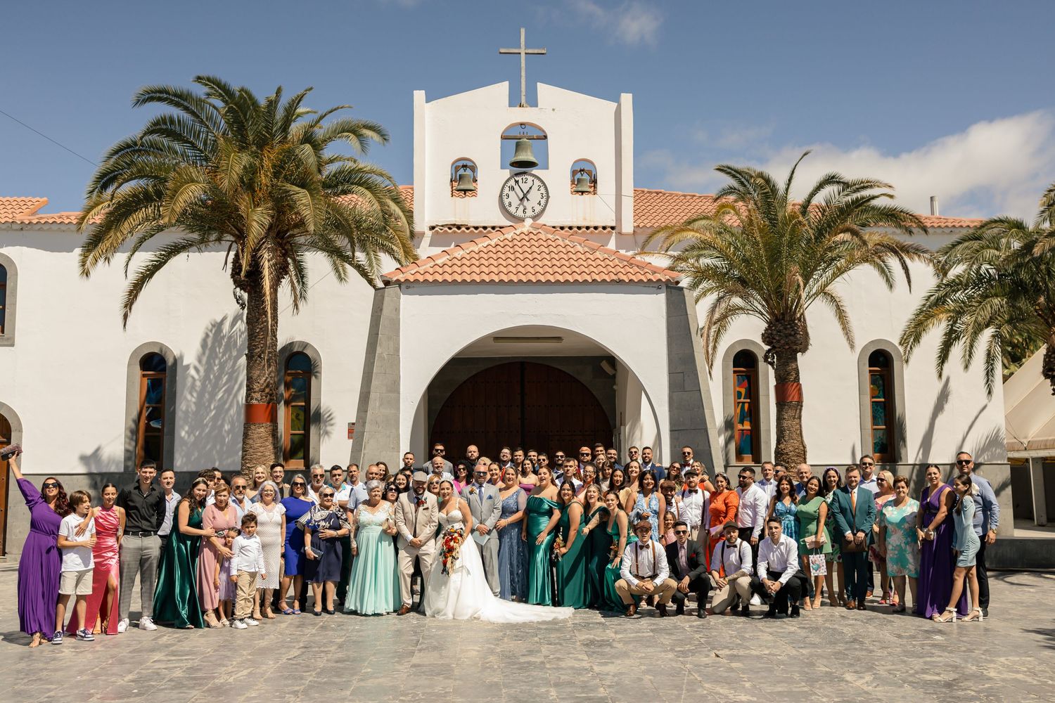 Panoramic view of wedding guests lined up outside Spanish-style church with palm trees.