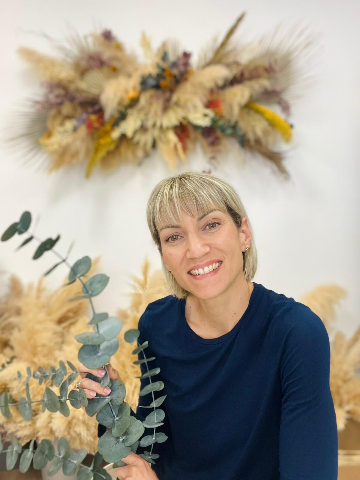 Four photos showing a person in a navy sweater posing with dried pampas grass and eucalyptus wall decorations.