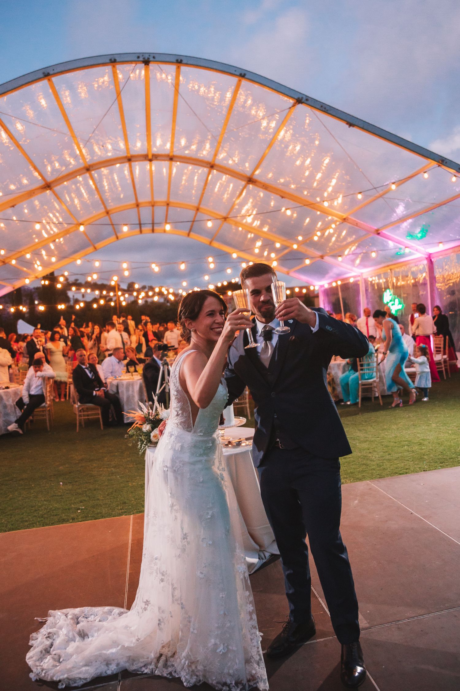 Wedding reception tent illuminated with string lights as couple dances on dance floor.