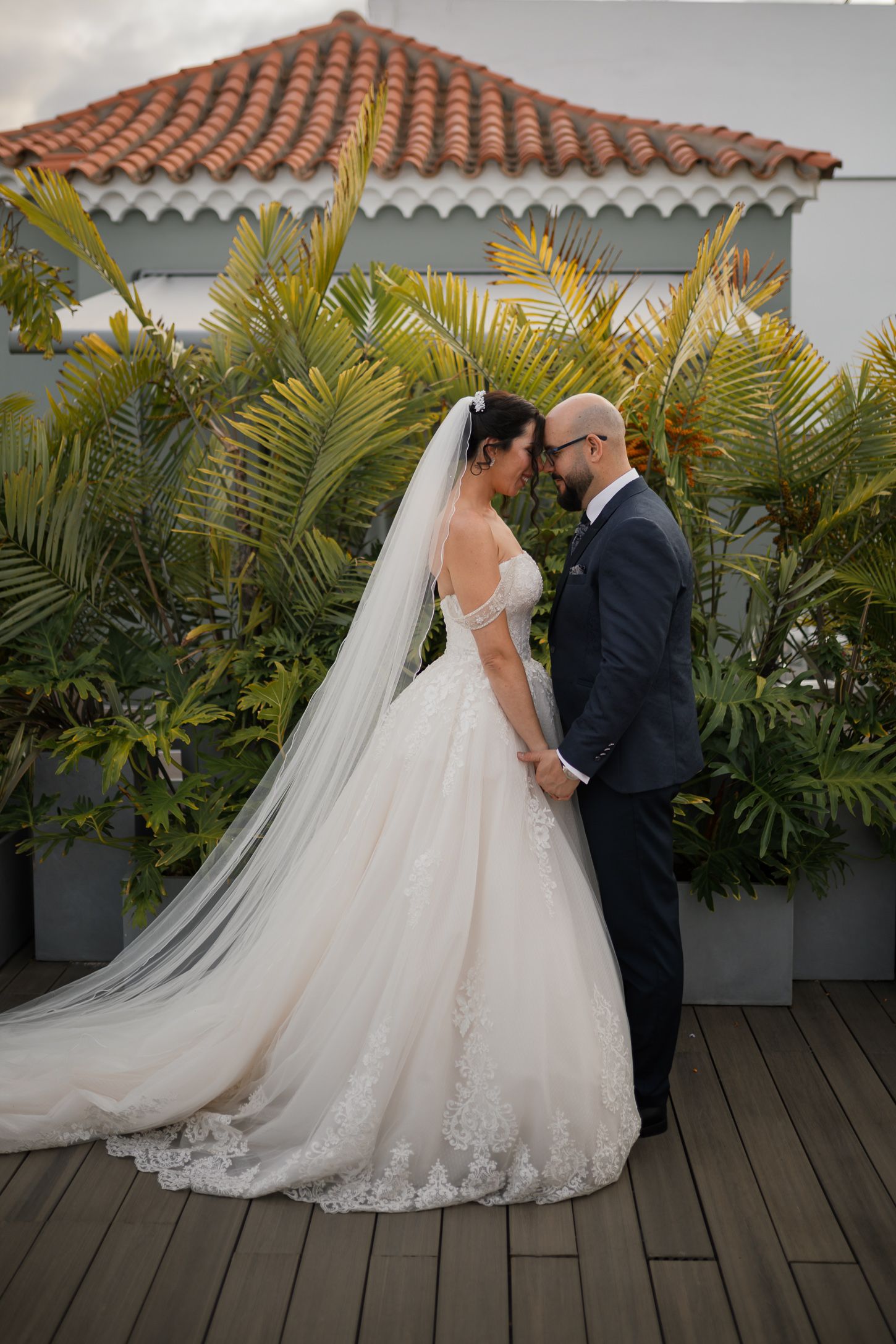 A wedding couple shares an intimate moment on a tropical terrace with palm fronds.