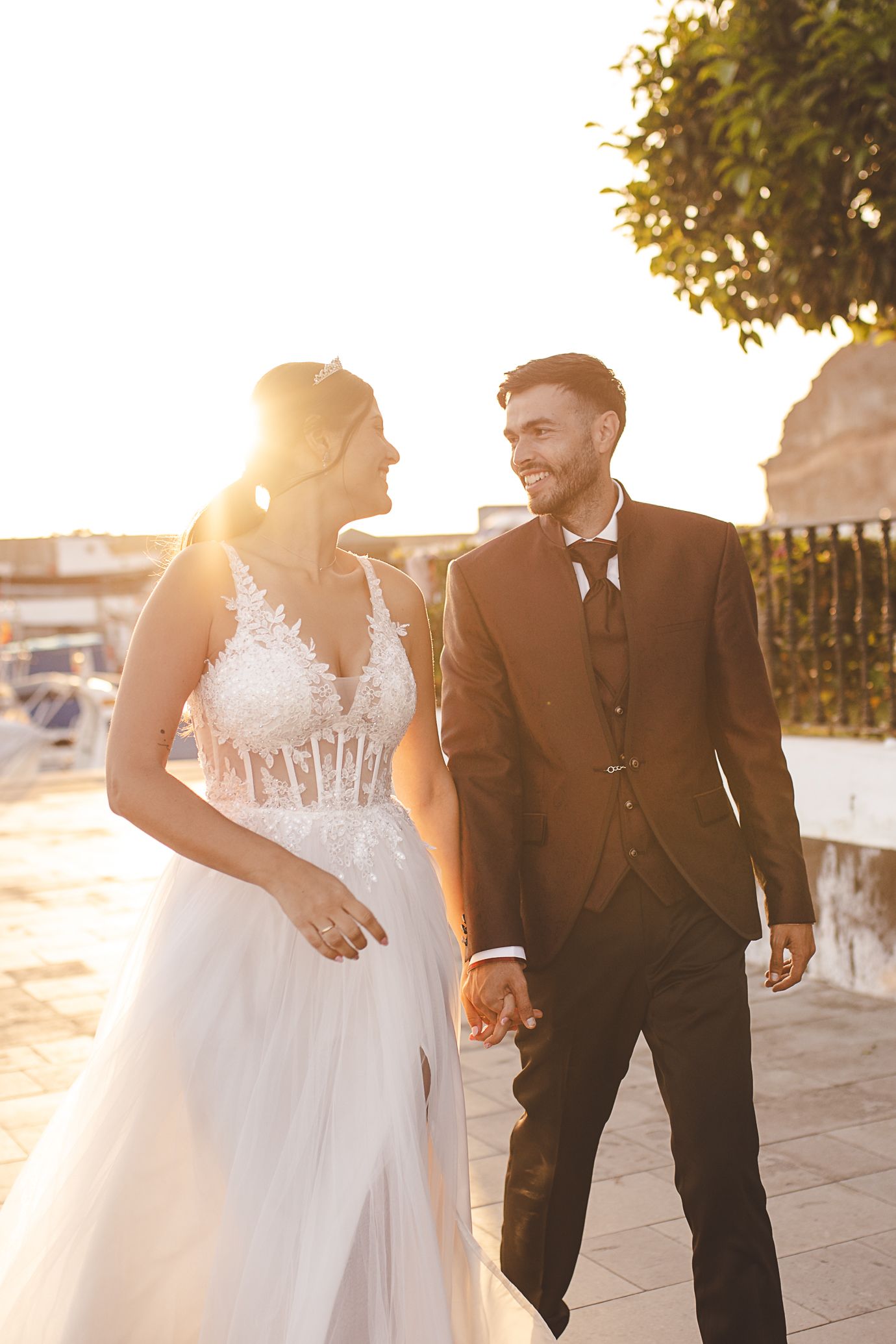 Newlywed couple holds hands while walking together at sunset, creating romantic silhouettes against golden light.