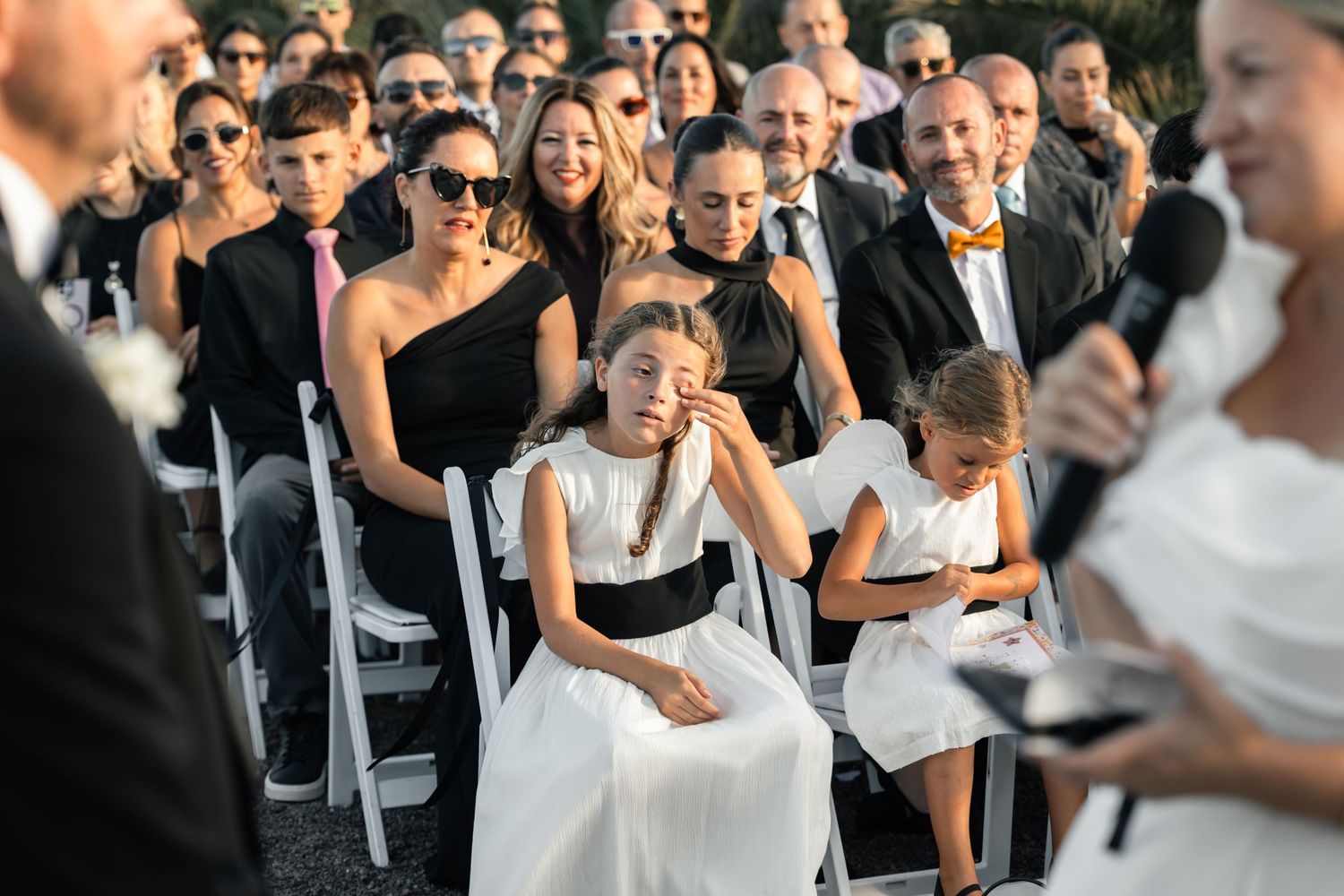 Emotional flower girl wiping tears at outdoor wedding ceremony with guests seated in white chairs.