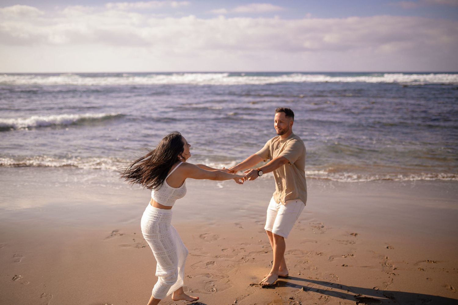 Two people playfully dancing and holding hands on a beach with waves in the background.