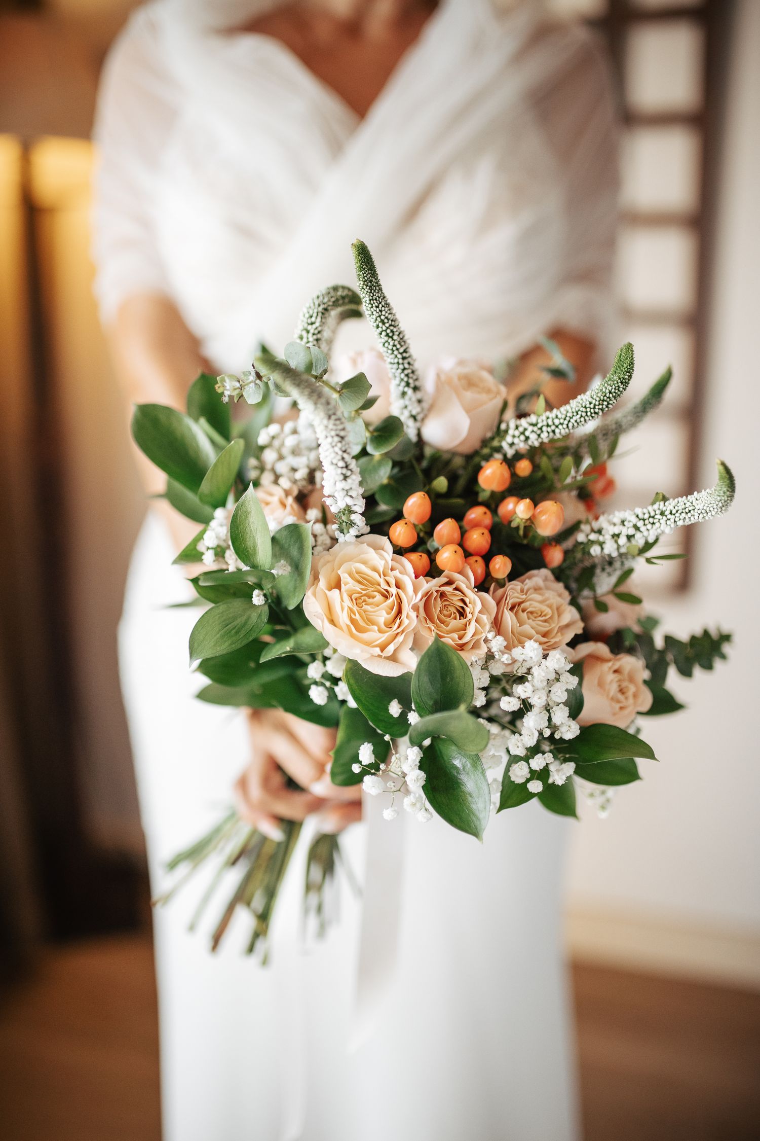Close-up of bridal bouquet featuring peach roses, white flowers and green foliage against white dress.