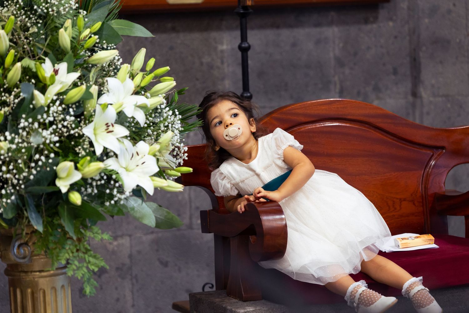 Young girl in white dress leaning on wooden church pew beside white lily flower arrangement.