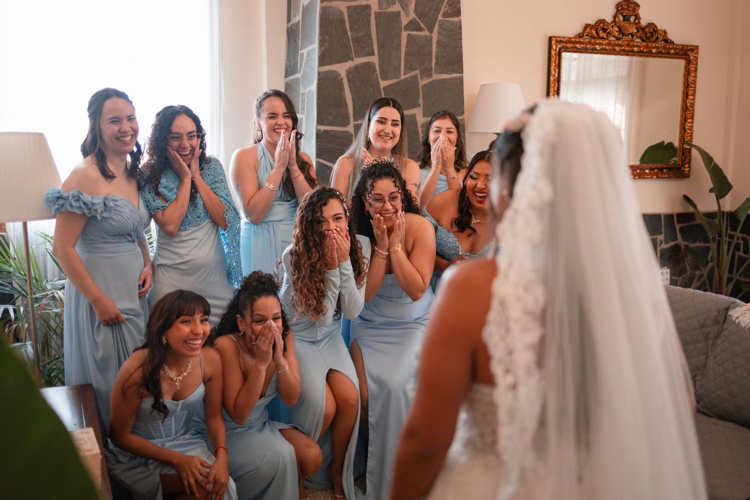 Bridesmaids in light blue dresses react with joy and tears seeing the bride in her wedding gown during an emotional first look.