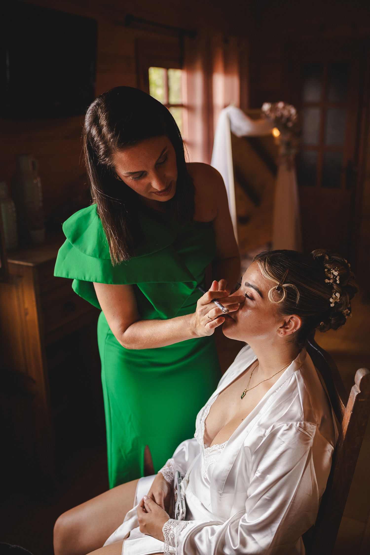 Makeup artist applies cosmetics to bride in white robe while wearing vibrant green dress in warm, dimly lit room.