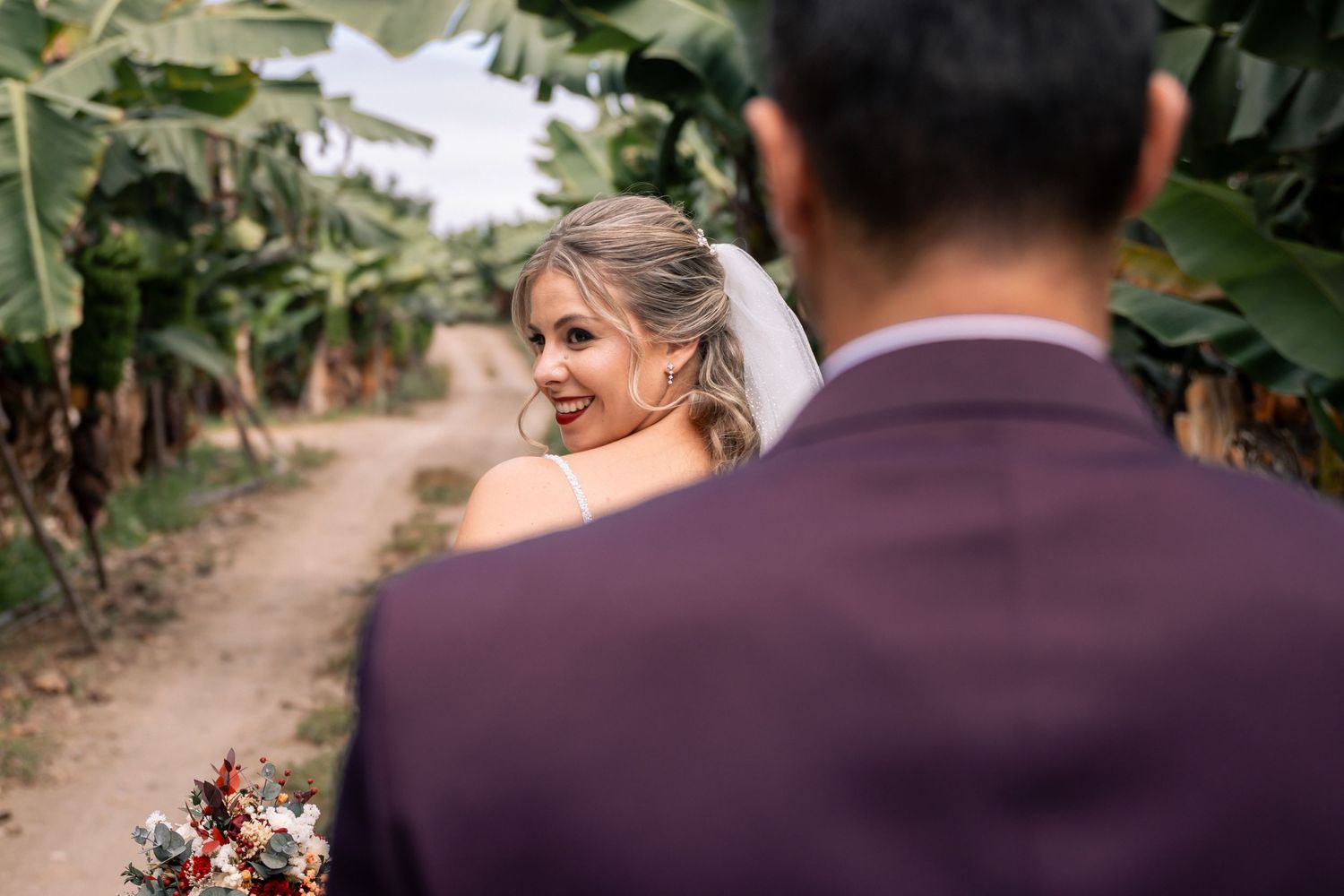 A couple walks down a rustic dirt path surrounded by lush greenery while holding hands.