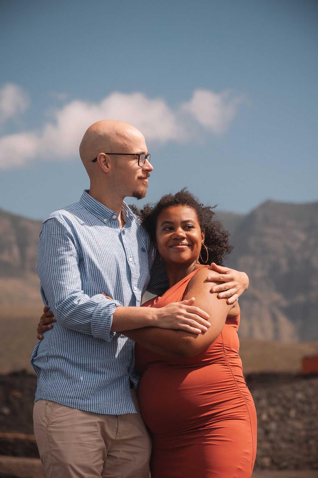 Loving couple shares tender moment during outdoor maternity photoshoot with mountains in background.