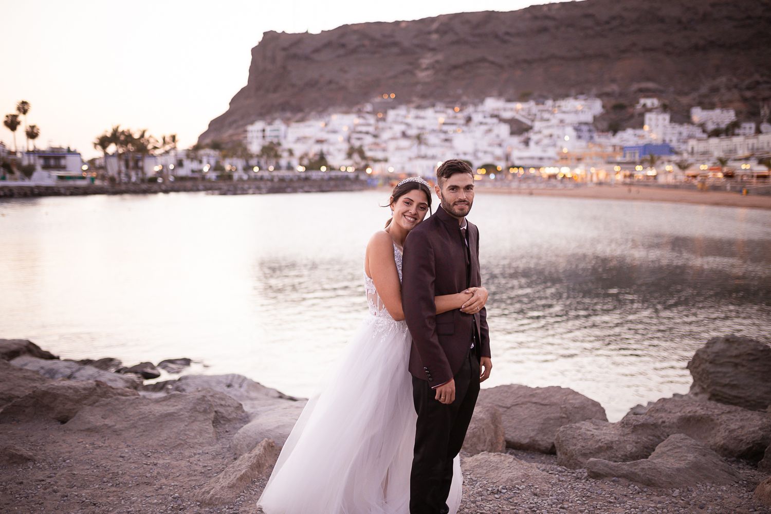 Wedding couple poses along rocky coastline with mountains and ocean in background.