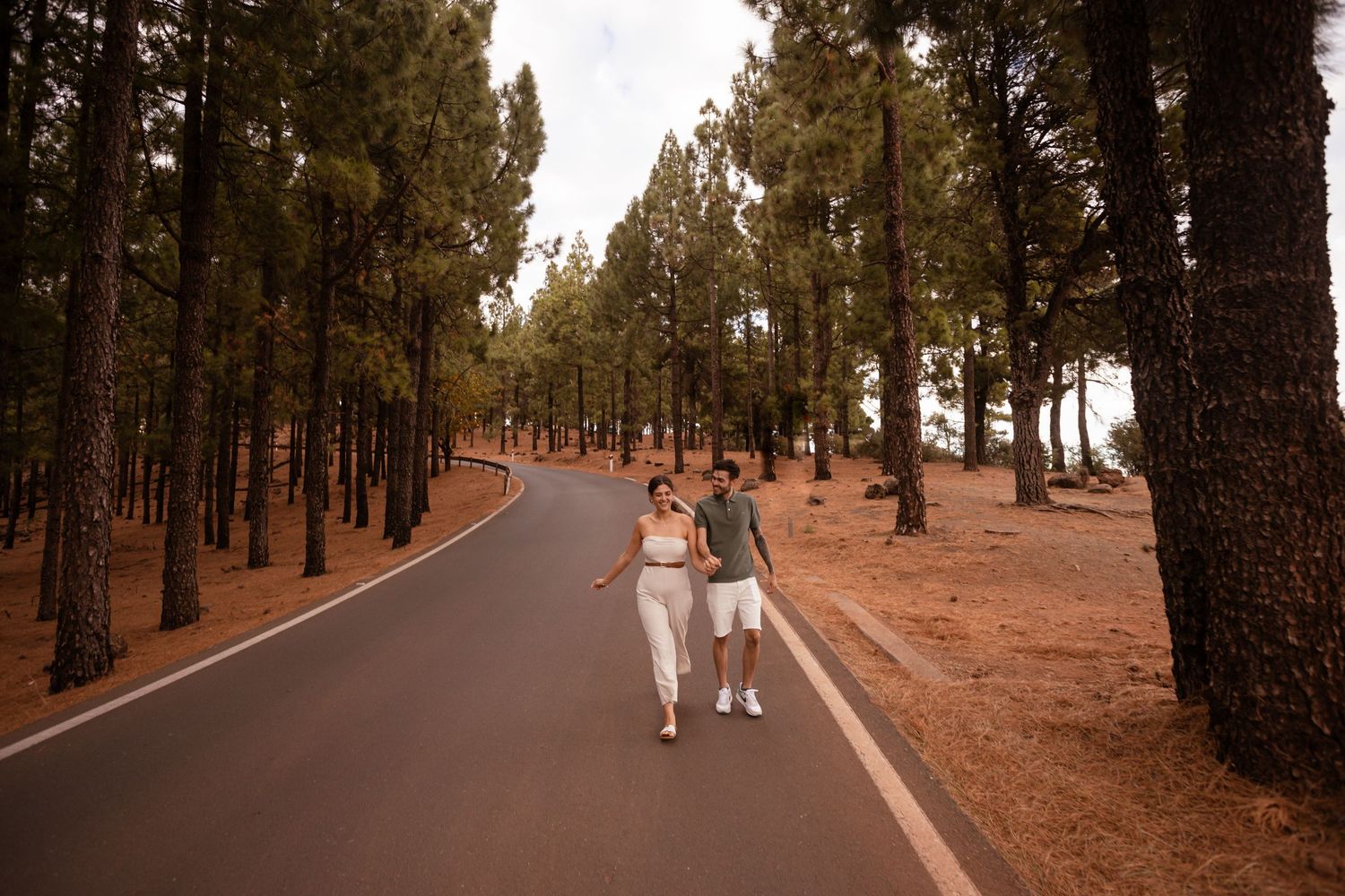 Couple walking hand in hand down a tree-lined road through a pine forest.