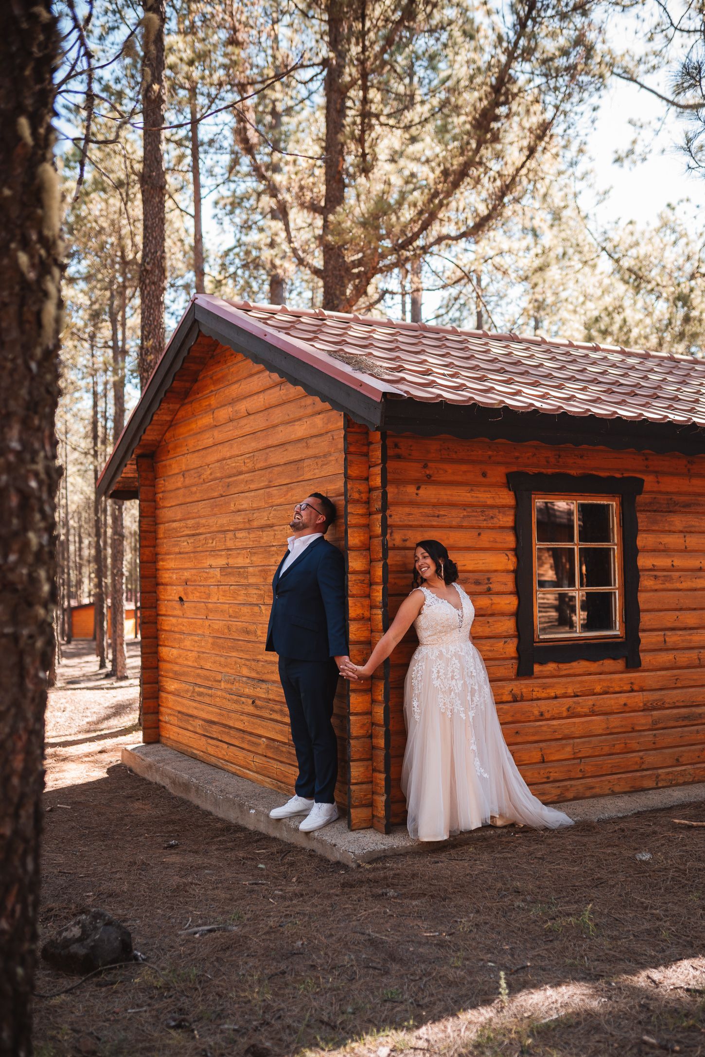 A romantic wedding couple poses outside a rustic wooden cabin in a sunlit forest setting.