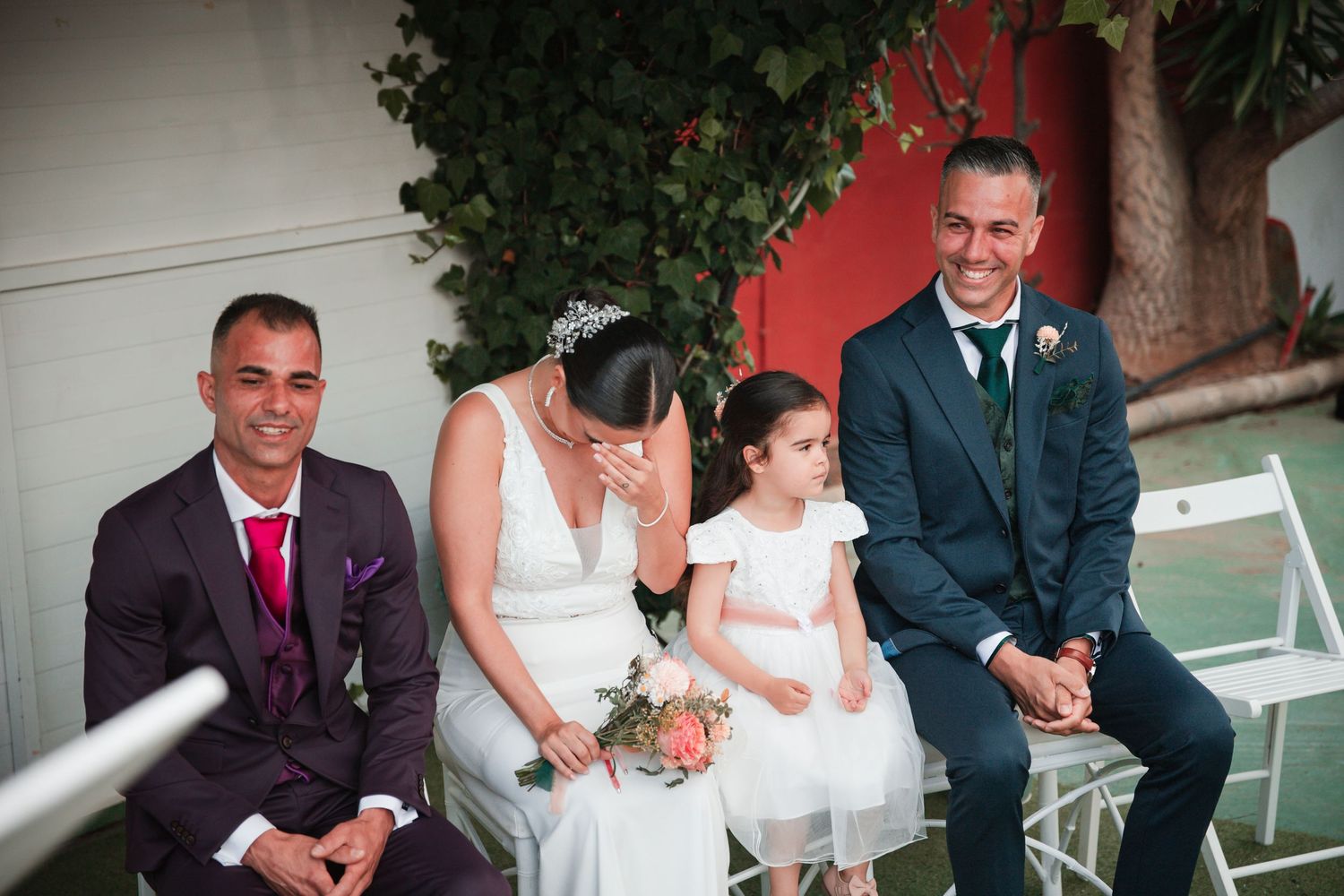 Wedding ceremony participants seated in formal attire outdoors.