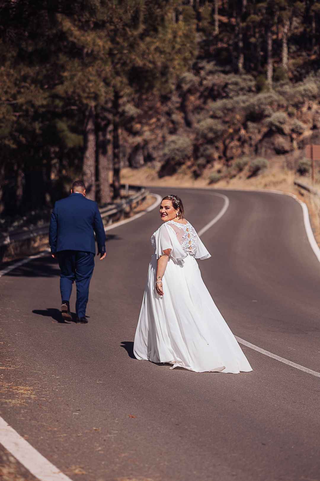 Wedding couple walking along a winding mountain road with pine trees in the background.