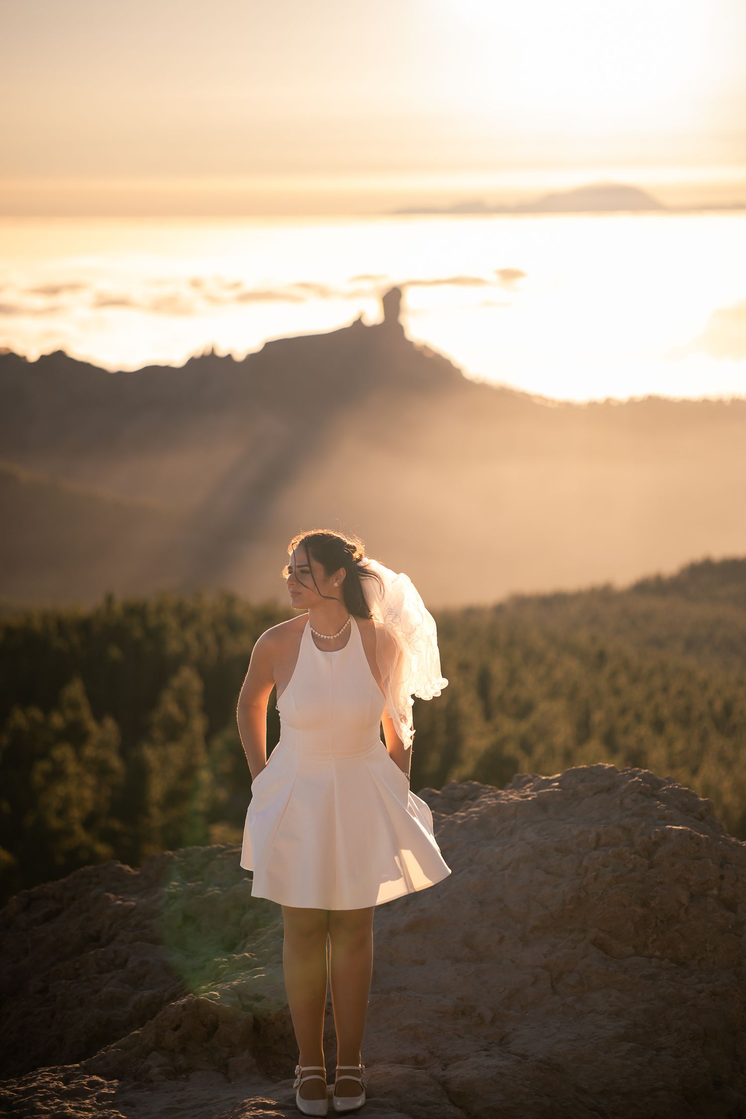 A person in white stands on a rocky overlook during golden hour with mountain views in the background.