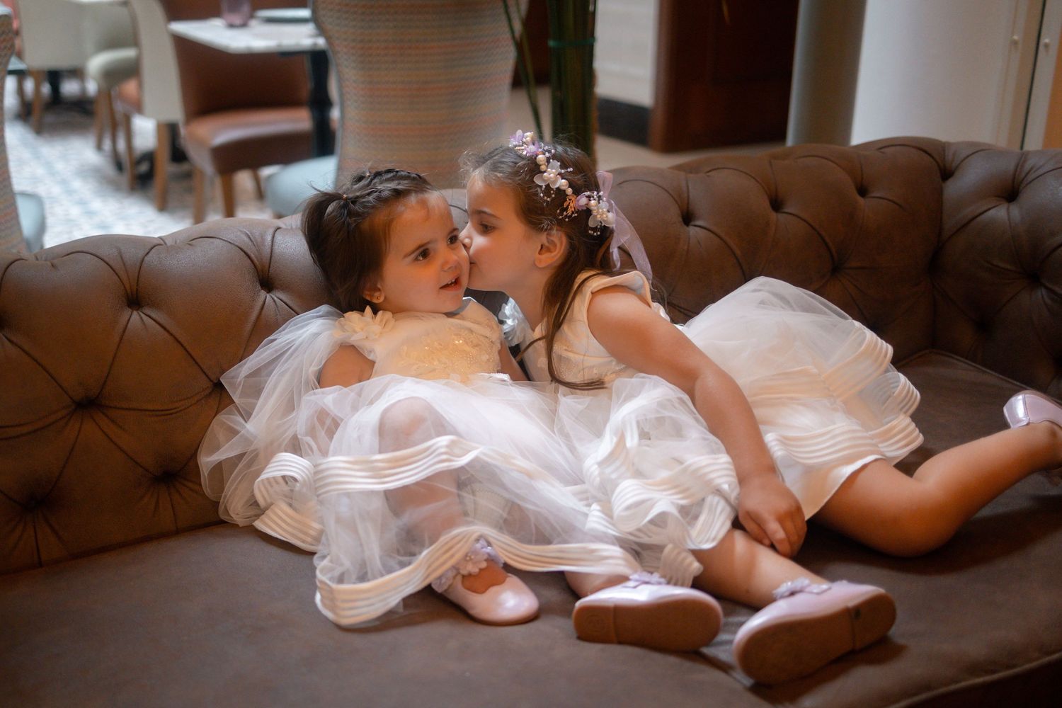 Two young flower girls in white tulle dresses sitting together on a brown tufted sofa, sharing a sweet moment.