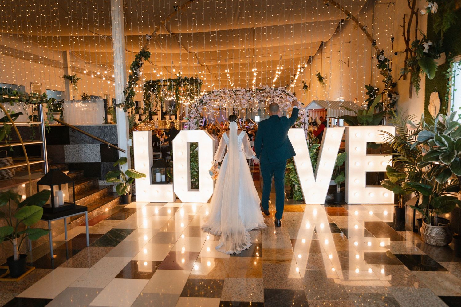 Illuminated LOVE marquee letters create a romantic ambiance at an indoor wedding reception with greenery and string lights.