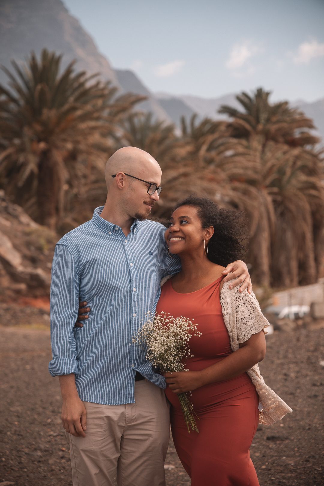 Couple poses for romantic photos among palm trees during sunset maternity session.