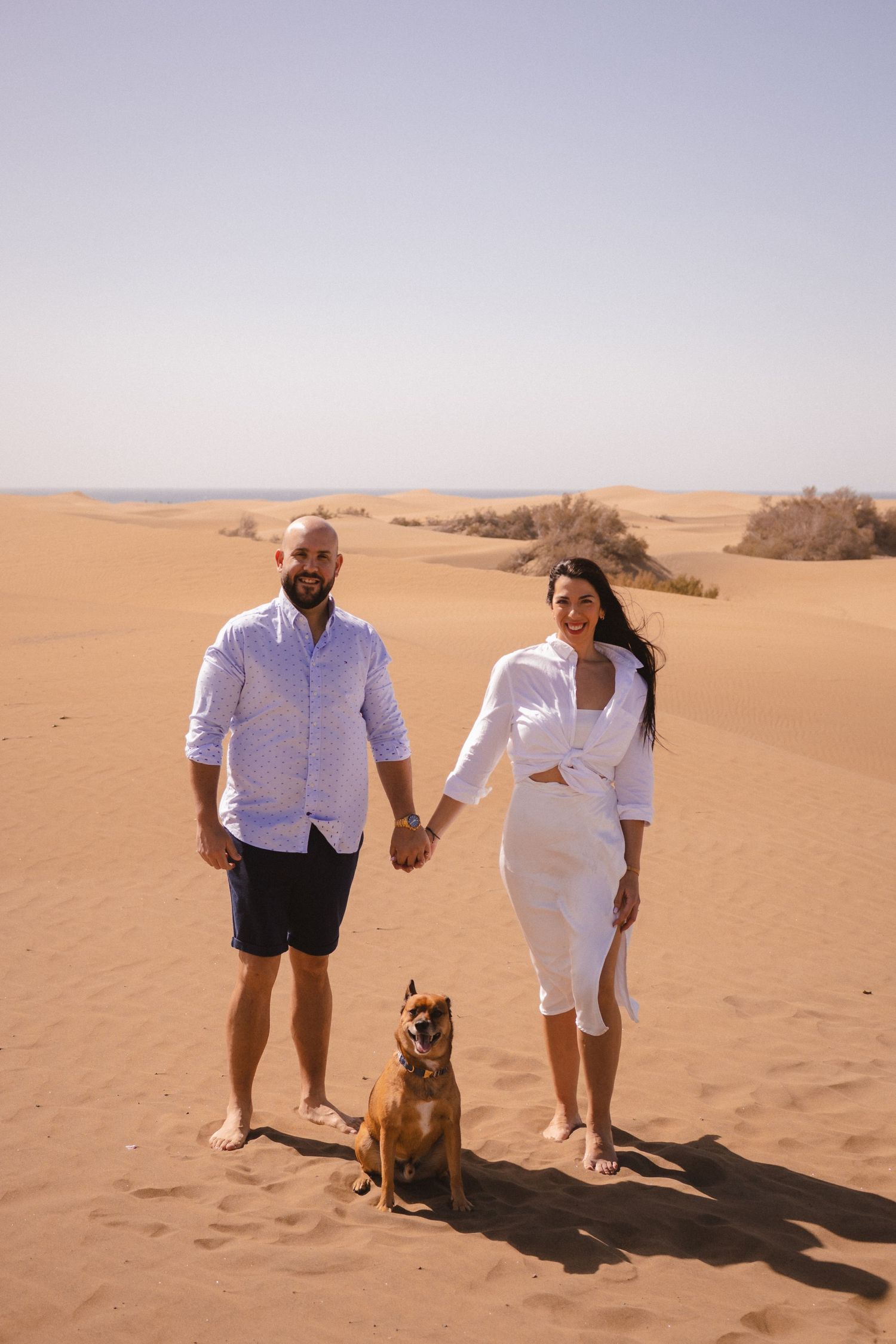 Couple in white outfits posing with their dog in desert sand dunes.