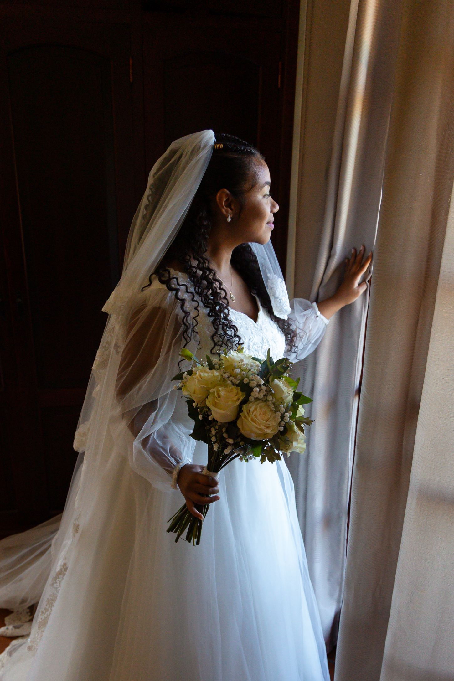 A bride in a white gown holds a yellow bouquet while looking out a sunlit window.
