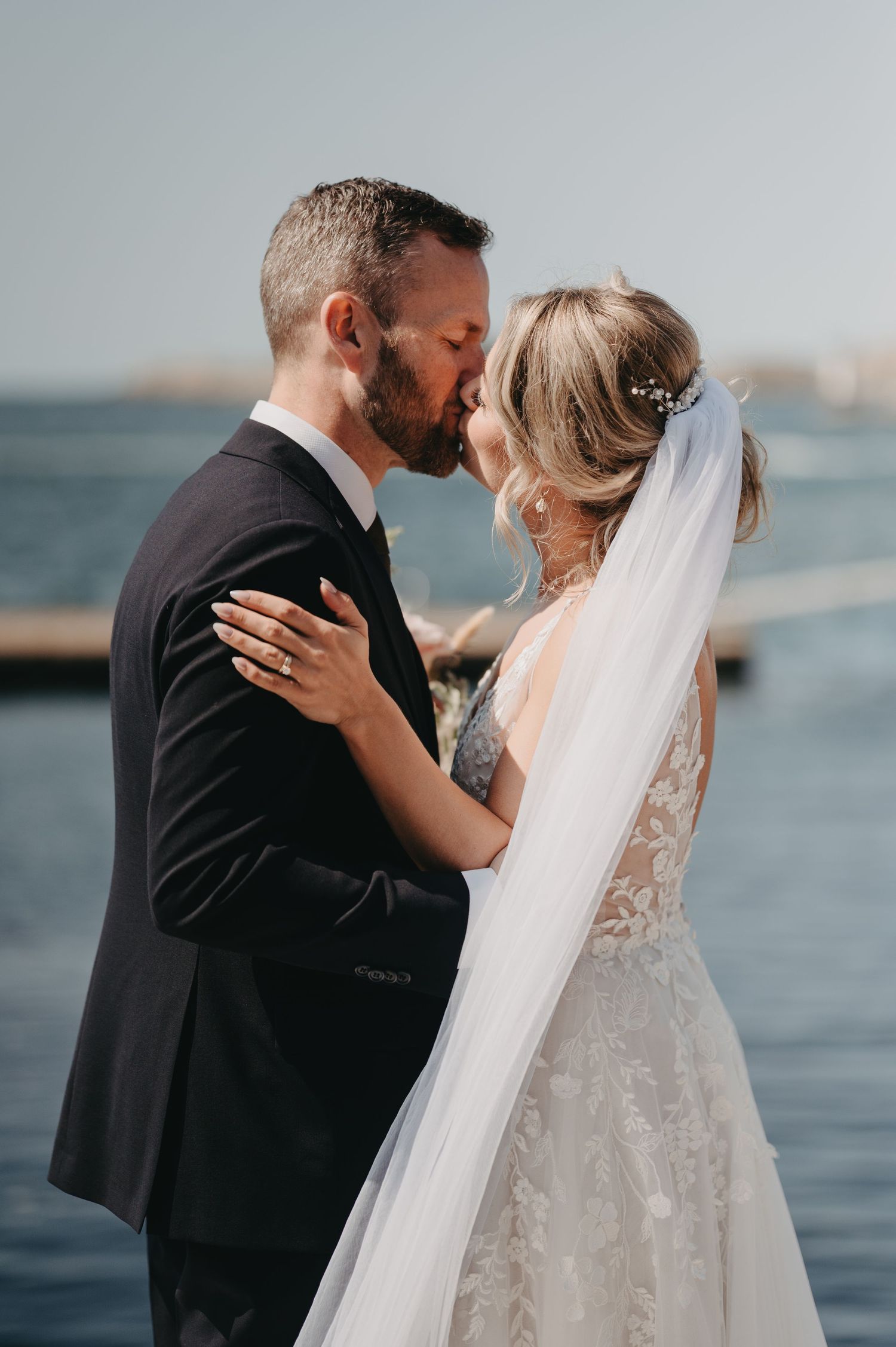 Bride and groom share a romantic kiss near the waterfront, bride wearing lace gown with veil and hair accessories.