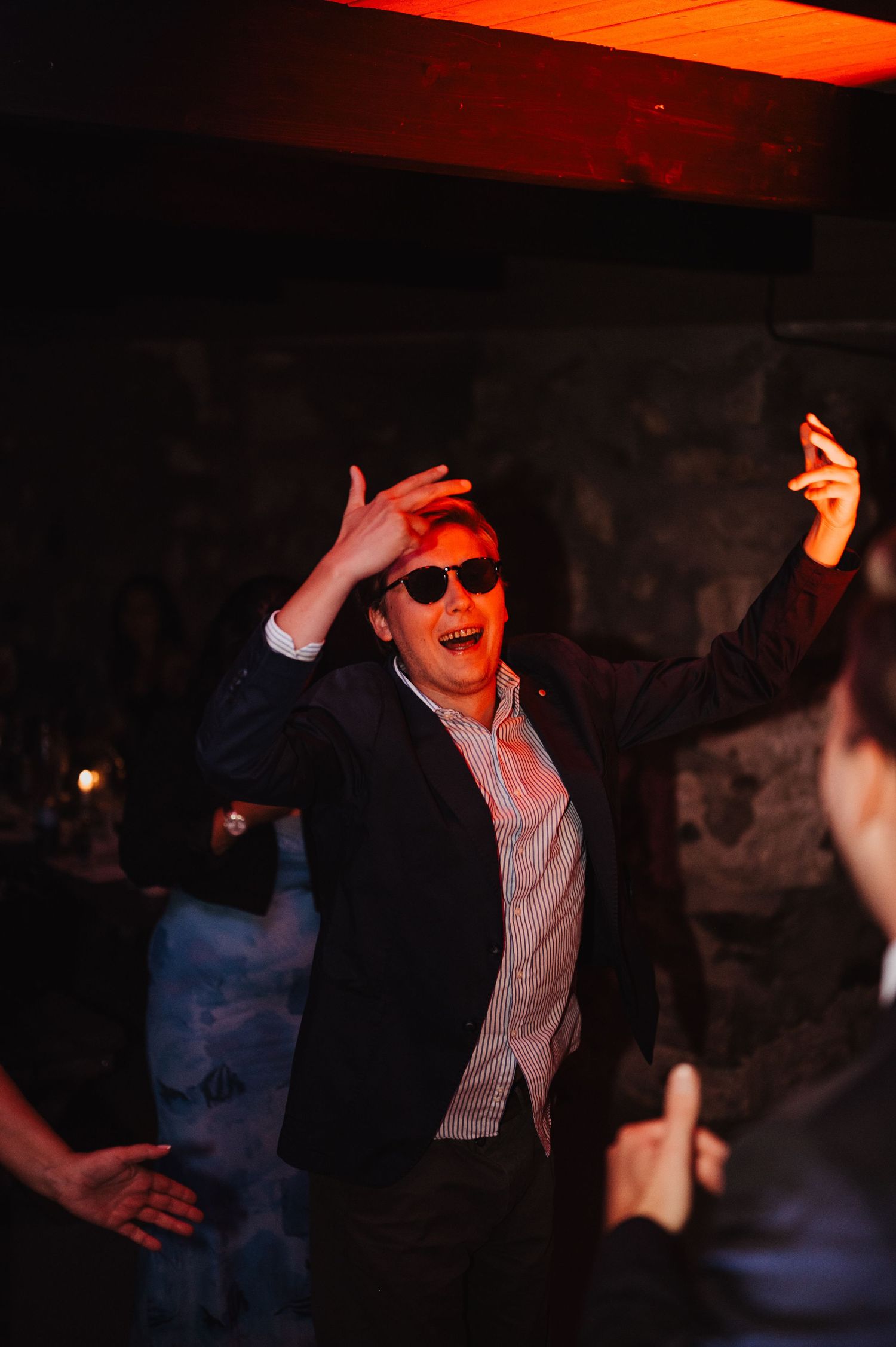 Energetic wedding guest dancing with sunglasses and arms raised under warm red lighting.