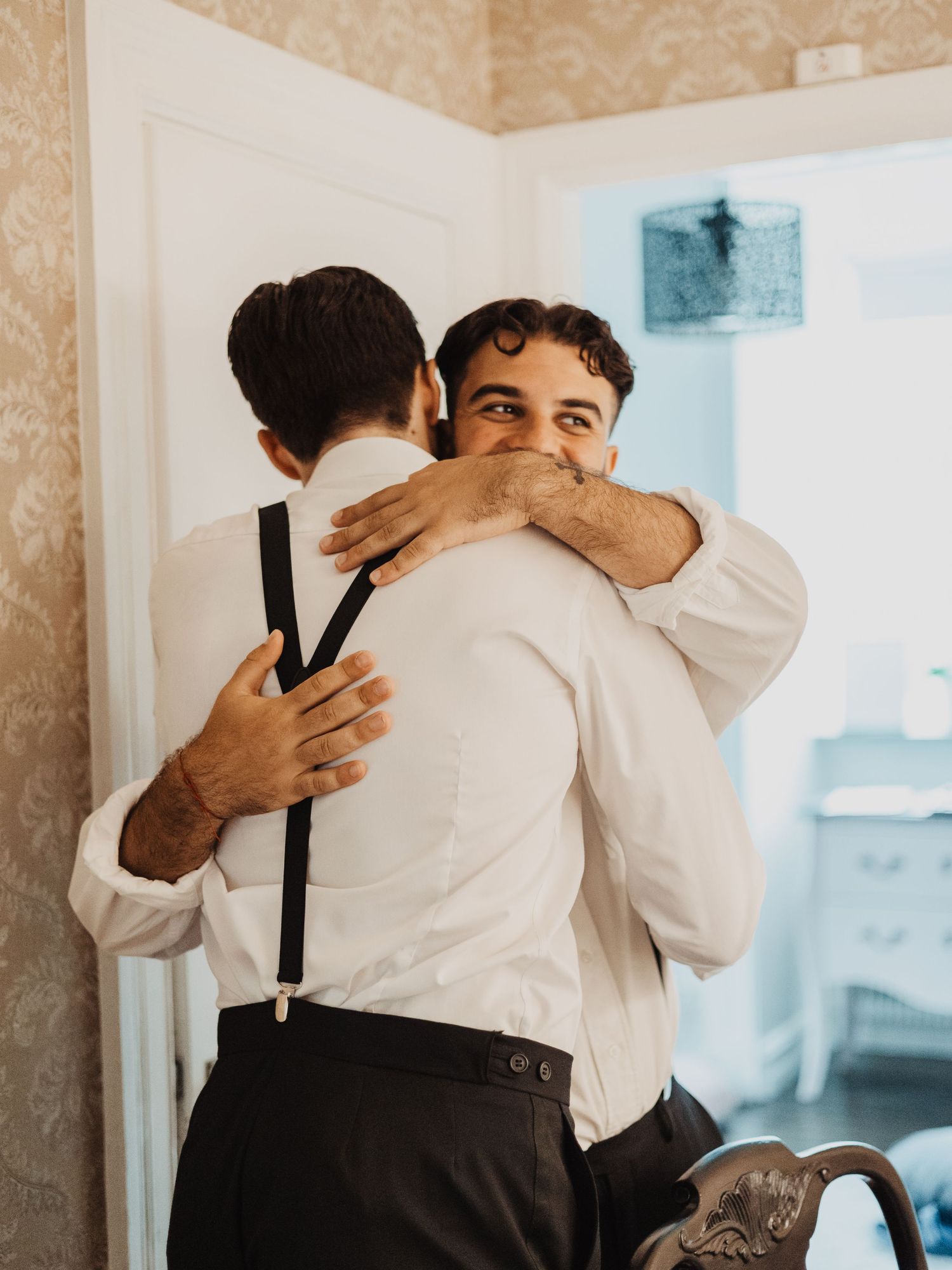 Two groomsmen sharing a warm embrace while getting dressed for a wedding celebration.