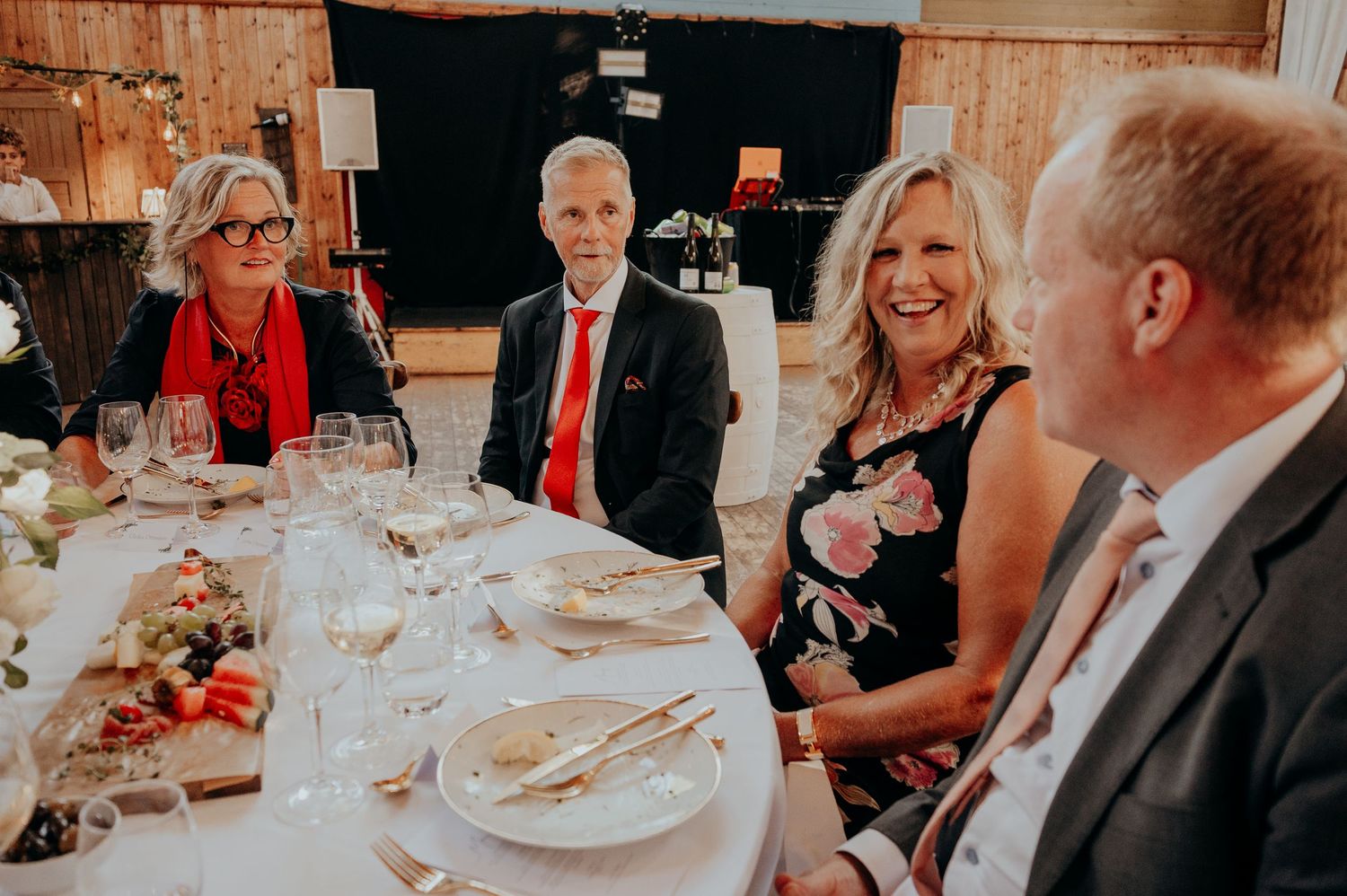Four elegantly dressed adults smiling and conversing at a formal dinner table set with wine glasses and floral centerpiece.