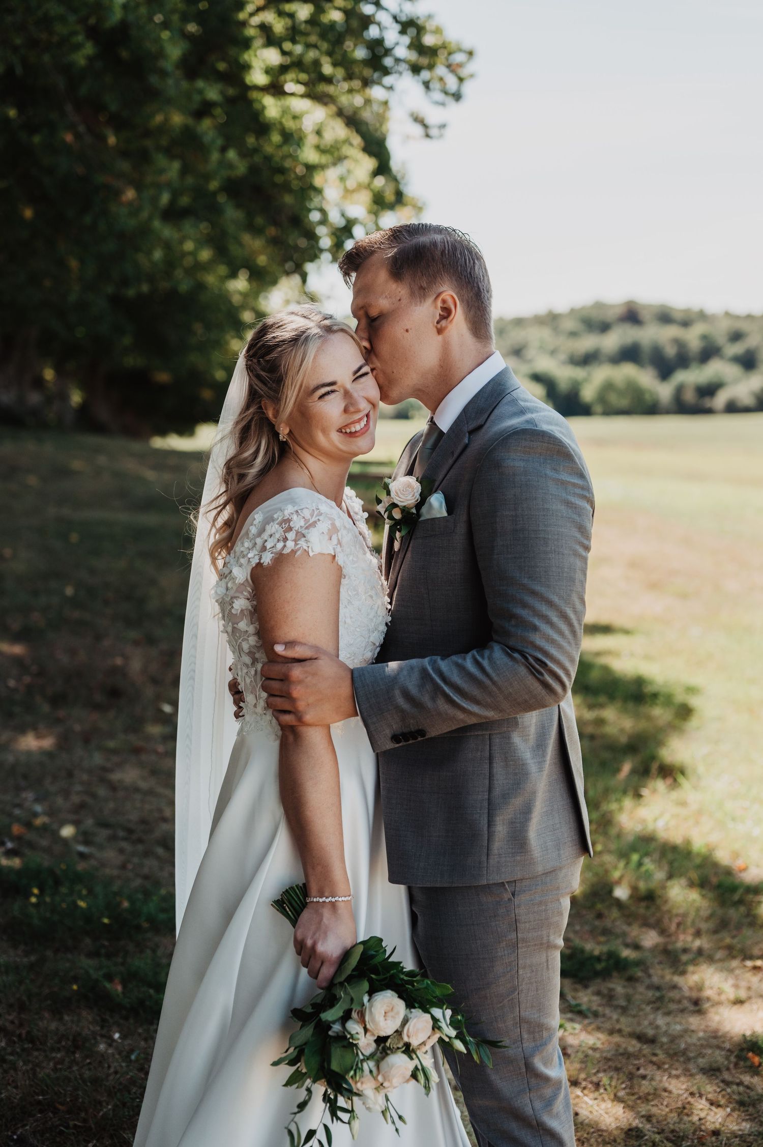 Smiling bride in lace gown and groom in gray suit share a romantic moment in a sunlit garden.