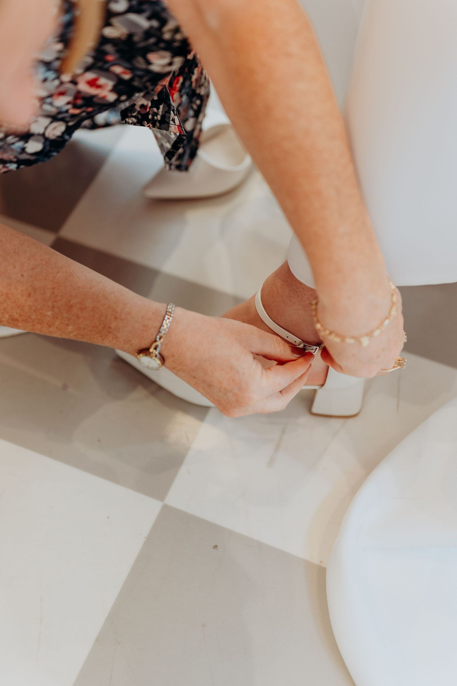 Close-up of hands fastening white block-heeled bridal shoe on brides foot on checkered salon floor.