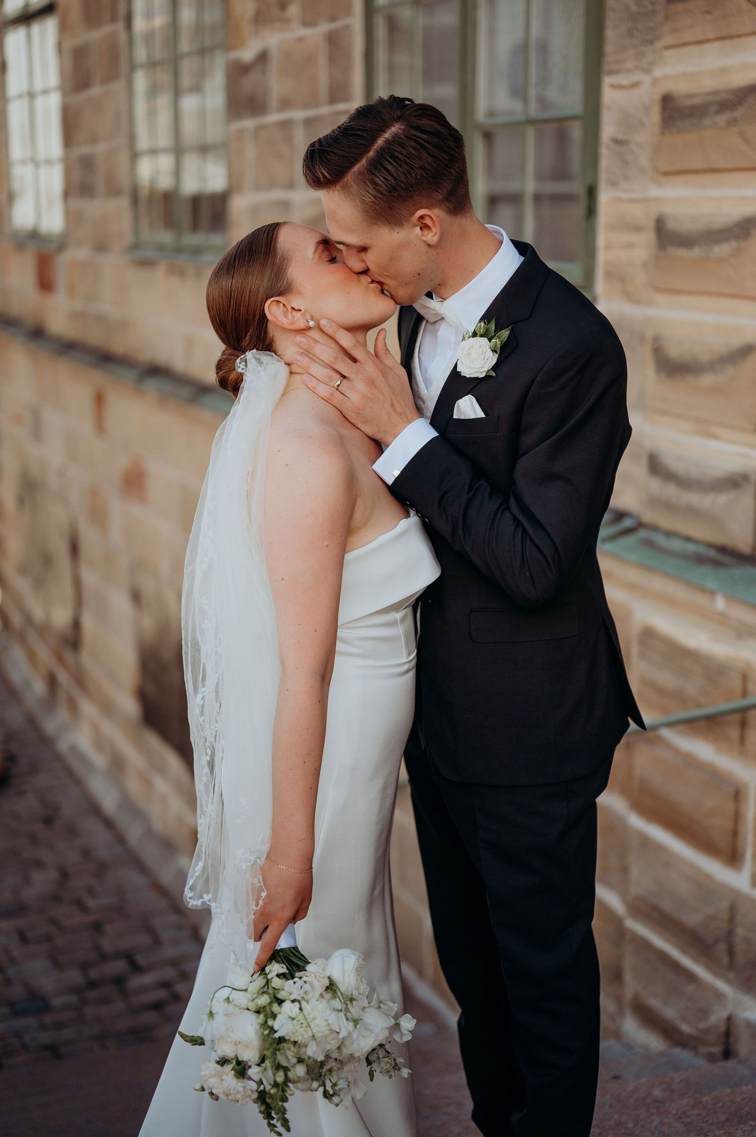 Bride and groom sharing a passionate kiss beside a historic sandstone building.