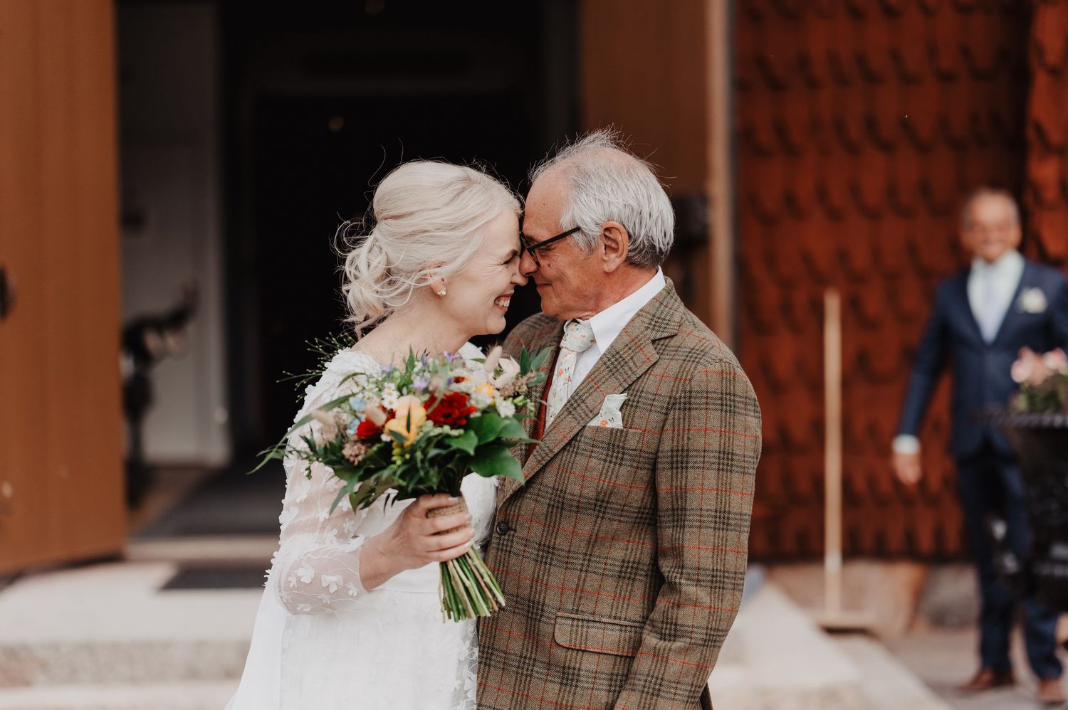 Bride and older man in plaid suit share tender forehead touch moment outdoors holding wedding bouquet.
