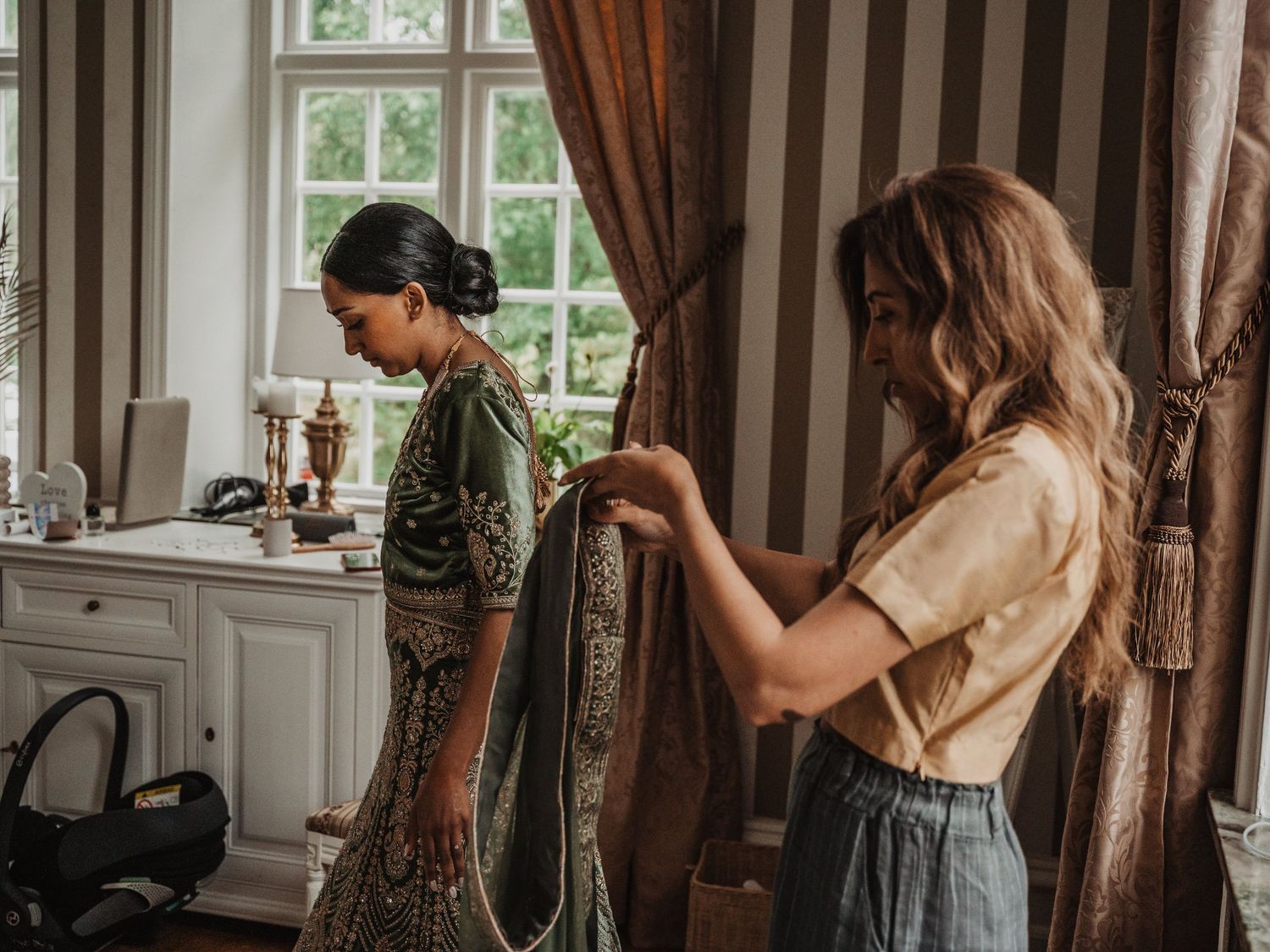 A mom helps a bride in a green embroidered lehenga get dressed in an elegant room.