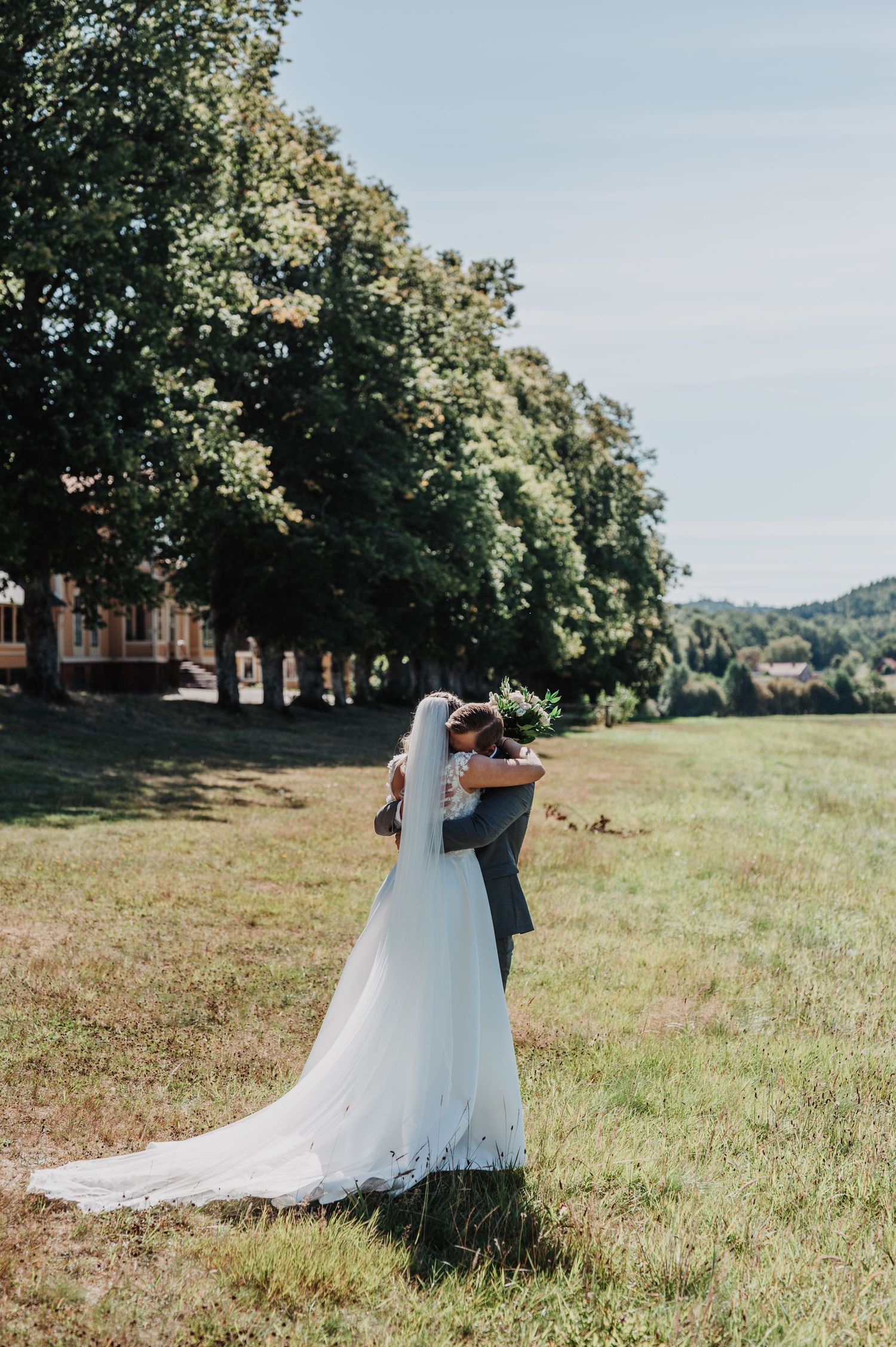 Bride and groom embracing in a sunlit meadow, bride wearing a flowing white gown with long veil, lush trees in background.