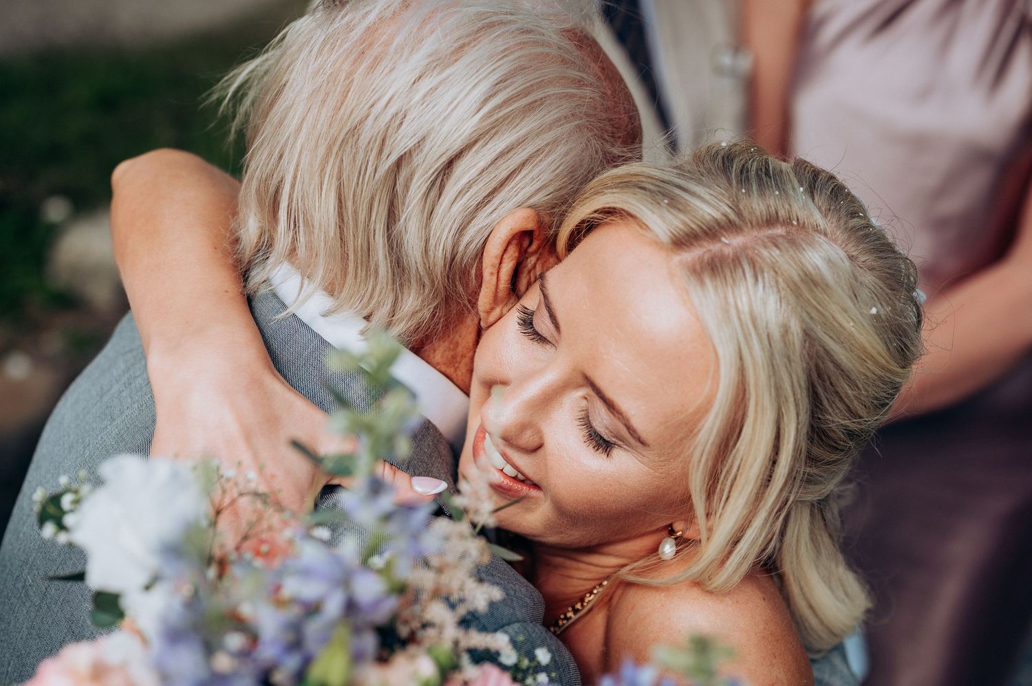 Blonde bride embraces older man tenderly while holding colorful wedding bouquet.
