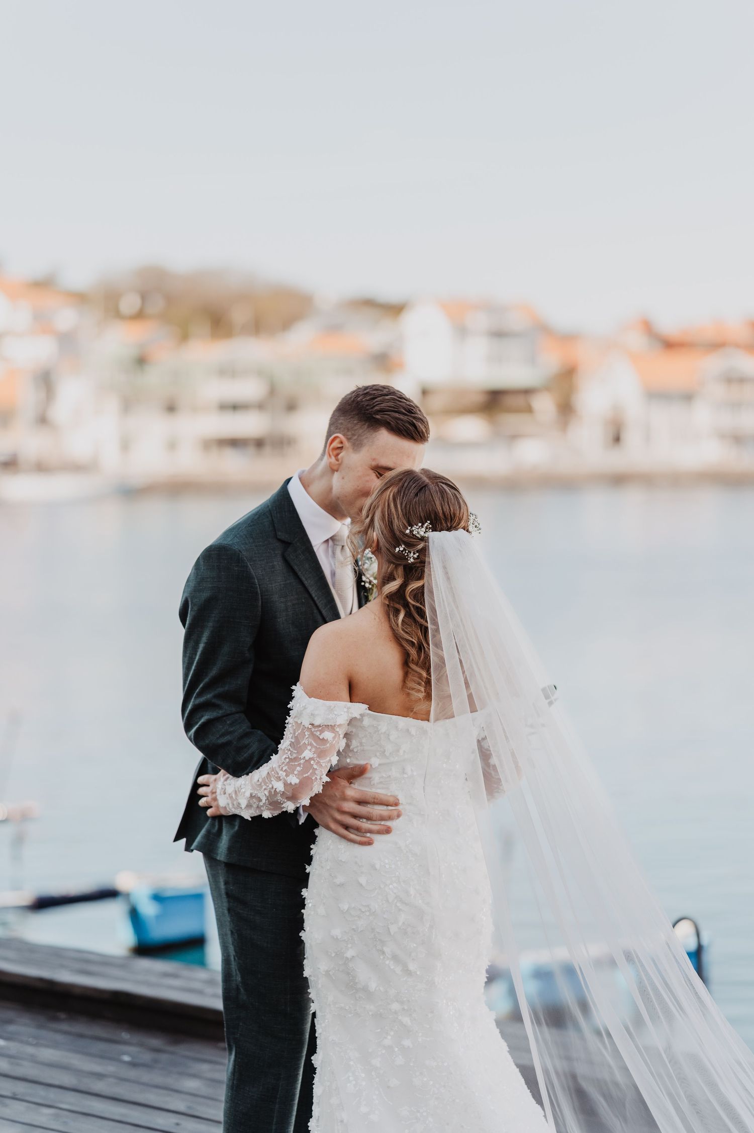 Bride and groom share a romantic kiss on a waterfront dock, bride in lace off-shoulder gown with veil.