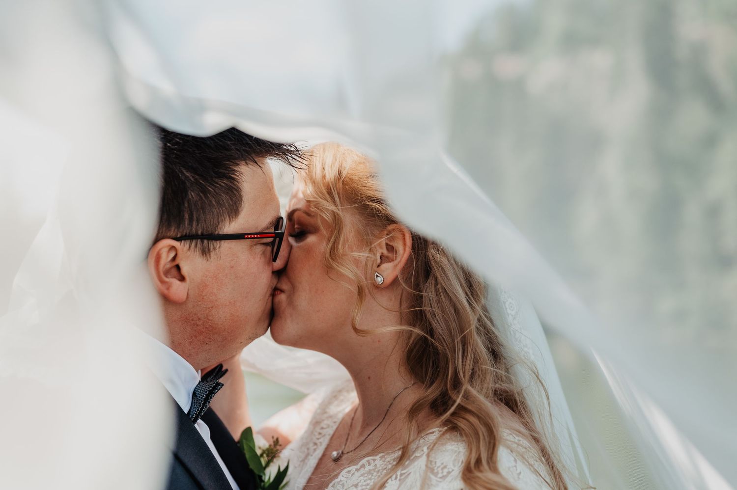 Bride and groom sharing romantic kiss beneath flowing white wedding veil.