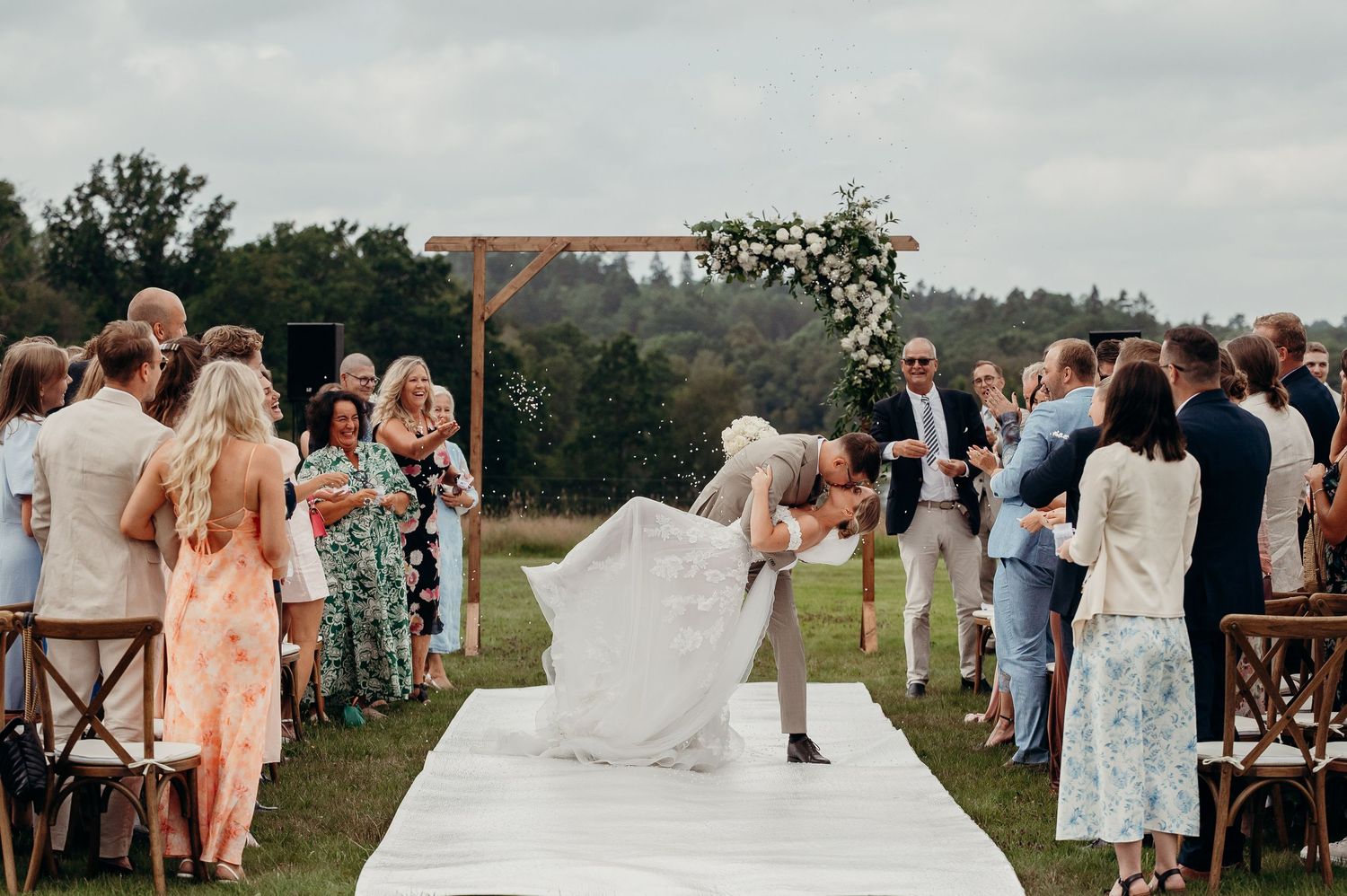 Groom dipping bride for a kiss at outdoor wedding ceremony on white aisle runner.