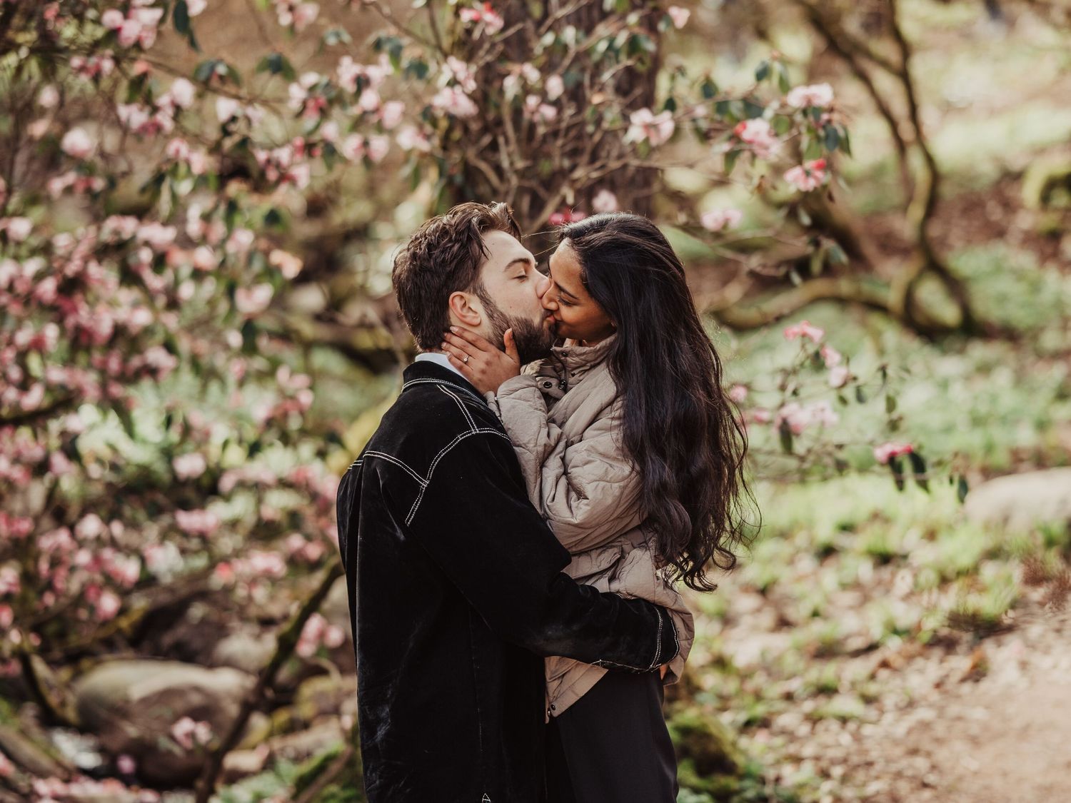 Romantic couple kissing passionately beneath blooming pink magnolia trees in a garden.