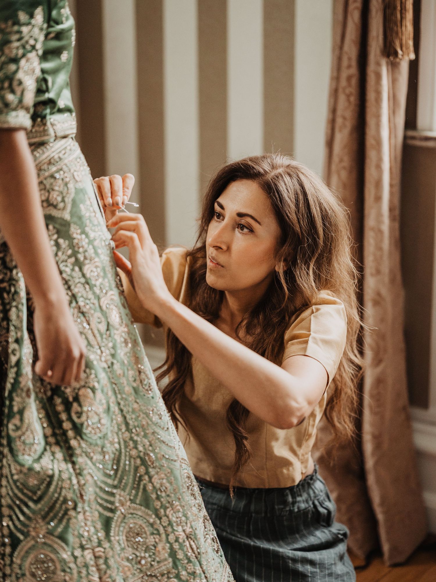 A focused mom kneeling to adjust the intricate embroidered skirt of a bridal lehenga.