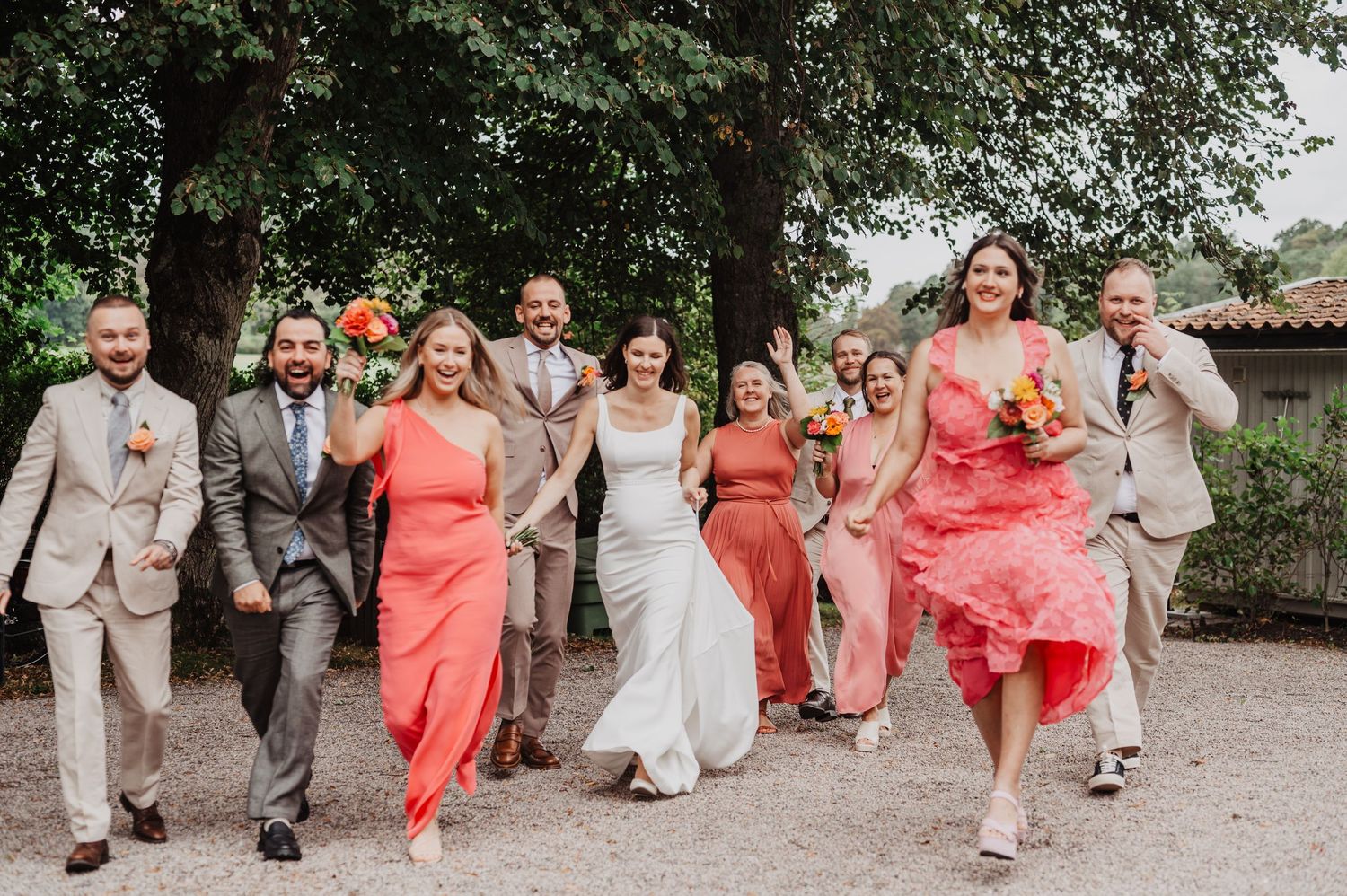 Wedding party in coral and beige attire walking and laughing together on a tree-lined path.