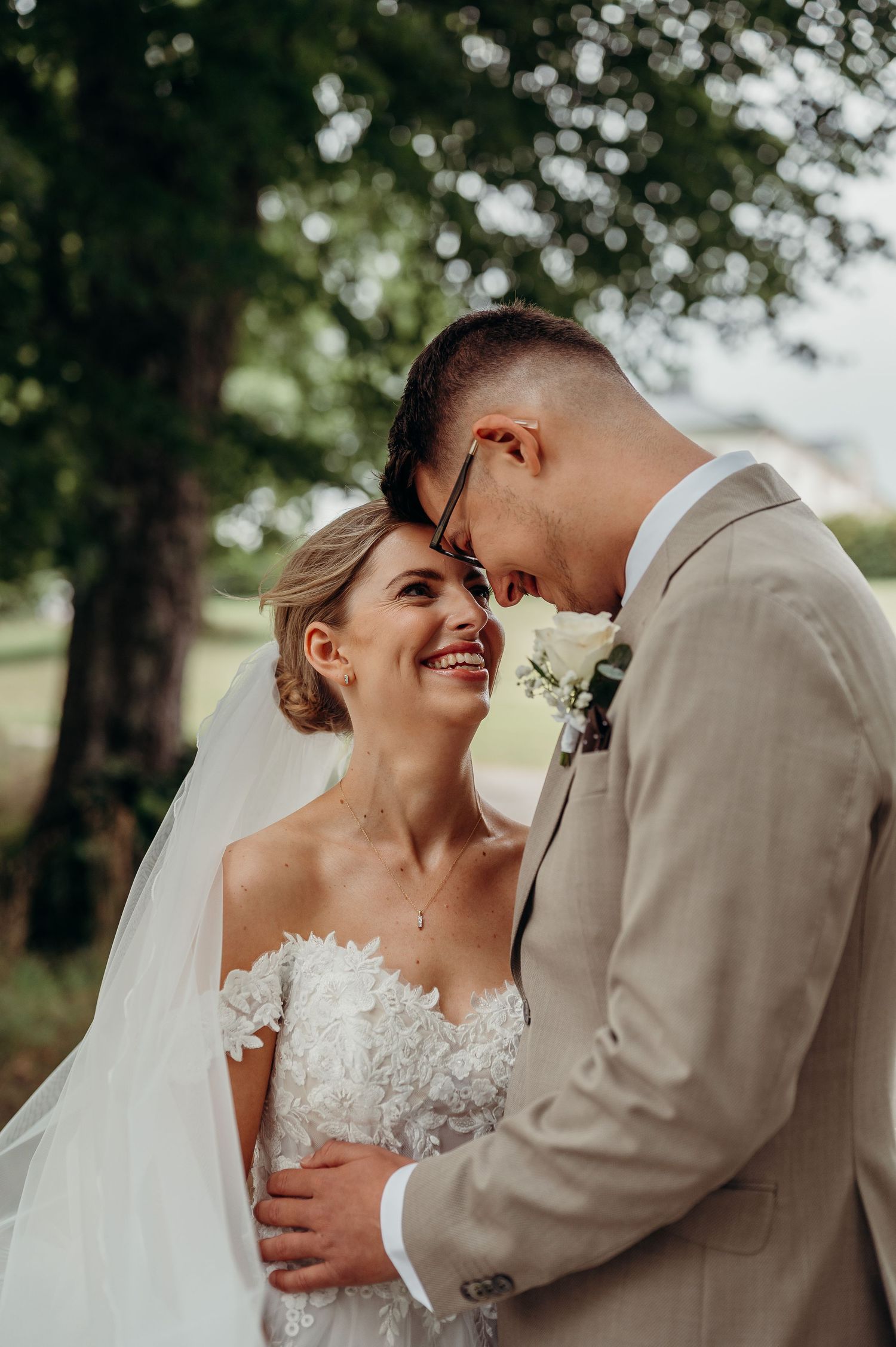 Bride and groom share an intimate moment, touching foreheads and smiling outdoors on their wedding day.