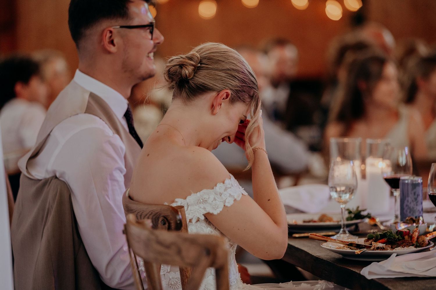 Bride laughing with head down beside groom at elegant wedding reception dinner.